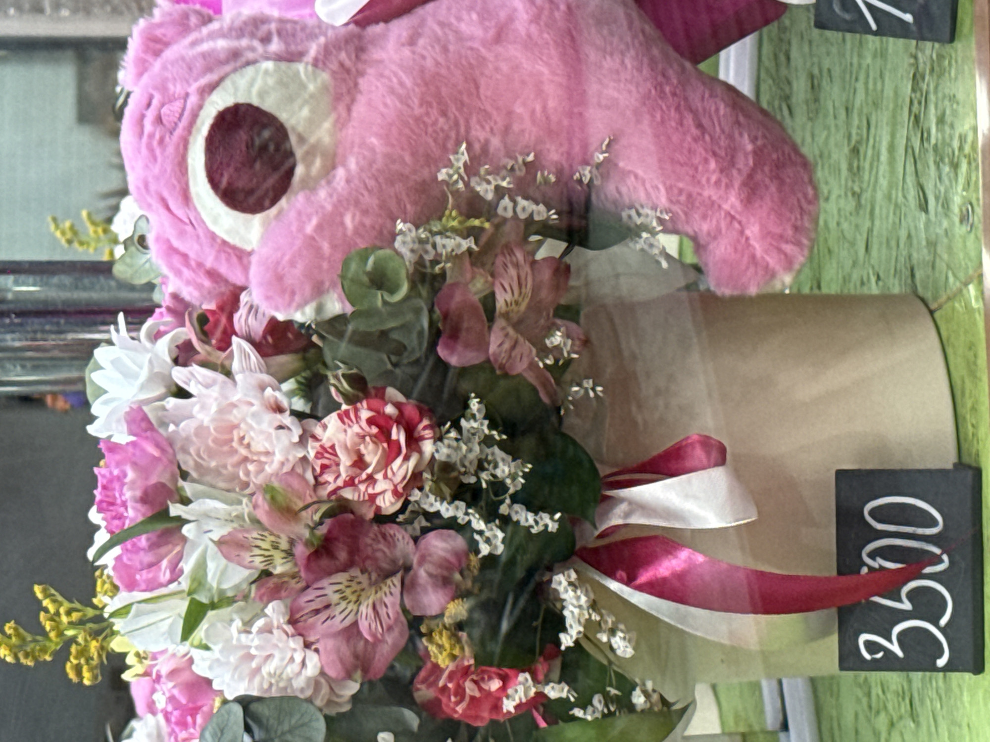 The image shows a bouquet of flowers and a pink stuffed animal. The bouquet contains a mix of white, pink, and red flowers, including what appears to be carnations and alstroemeria. The flowers are arranged with greenery and small white flowers. The bouquet is wrapped in brown paper. Above the bouquet is a pink stuffed animal with one large eye. The bouquet is tied with a white and pink ribbon. A small black sign with the number "3500" written in white is visible.
