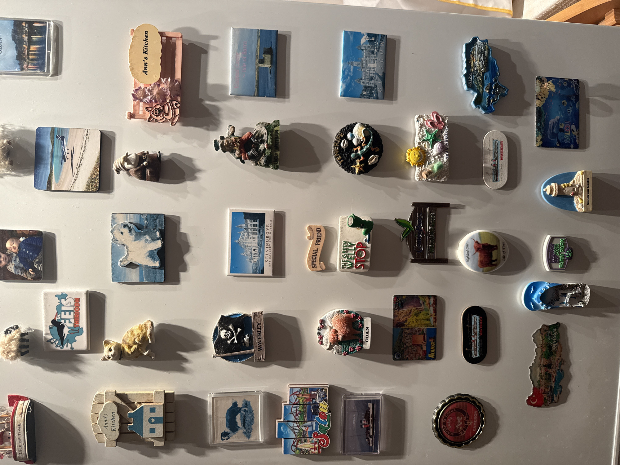 The image shows a collection of magnets on a refrigerator door. The magnets are of varying sizes, shapes, and designs. Some appear to be souvenirs from different places, including "Isle of Barra," "Glasgow," "Oban," "Antalya," "Alanya," "Kelvingrove Art Gallery and Museum," and "Salou." There are also magnets with images of a highland cow, a pirate skull and crossbones, and a green welly stop. Some of the magnets have text on them, such as "Ann's Kitchen," "Special Friend," "Sundrum Castle," "Farm Park," and "Isle of Tiree."
