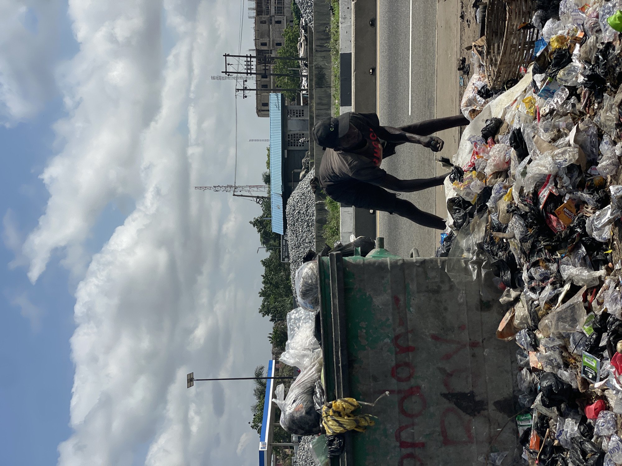 In Isunba, Nigeria, two individuals are scavenging through a large pile of trash beside a road. The trash pile contains various plastic bags and discarded items. A green dumpster is partially visible, filled with more trash. The environment includes power lines, a blue building, and a gas station in the background. The sky is cloudy.