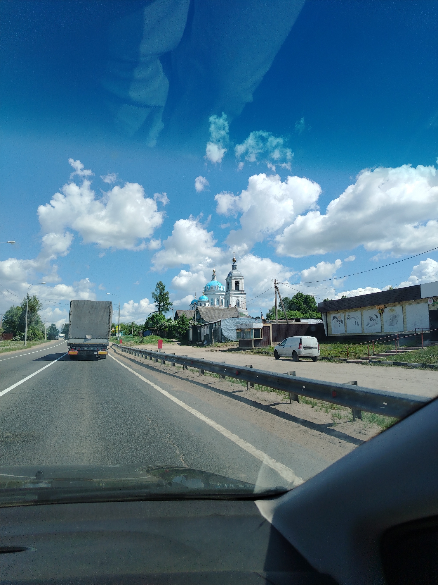 A photograph taken from inside a vehicle shows a view of a highway in Новое, Russia. A large truck is traveling in the same direction in the left lane. On the right side of the road, a gray metal guardrail runs along the shoulder. Beyond the guardrail, a white van is parked near a one-story building with drawings of animals on its exterior. In the background, a Russian Orthodox church with blue domes and white walls stands among trees and houses. The sky is blue with scattered white clouds. Reflections are visible on the windshield of the car.