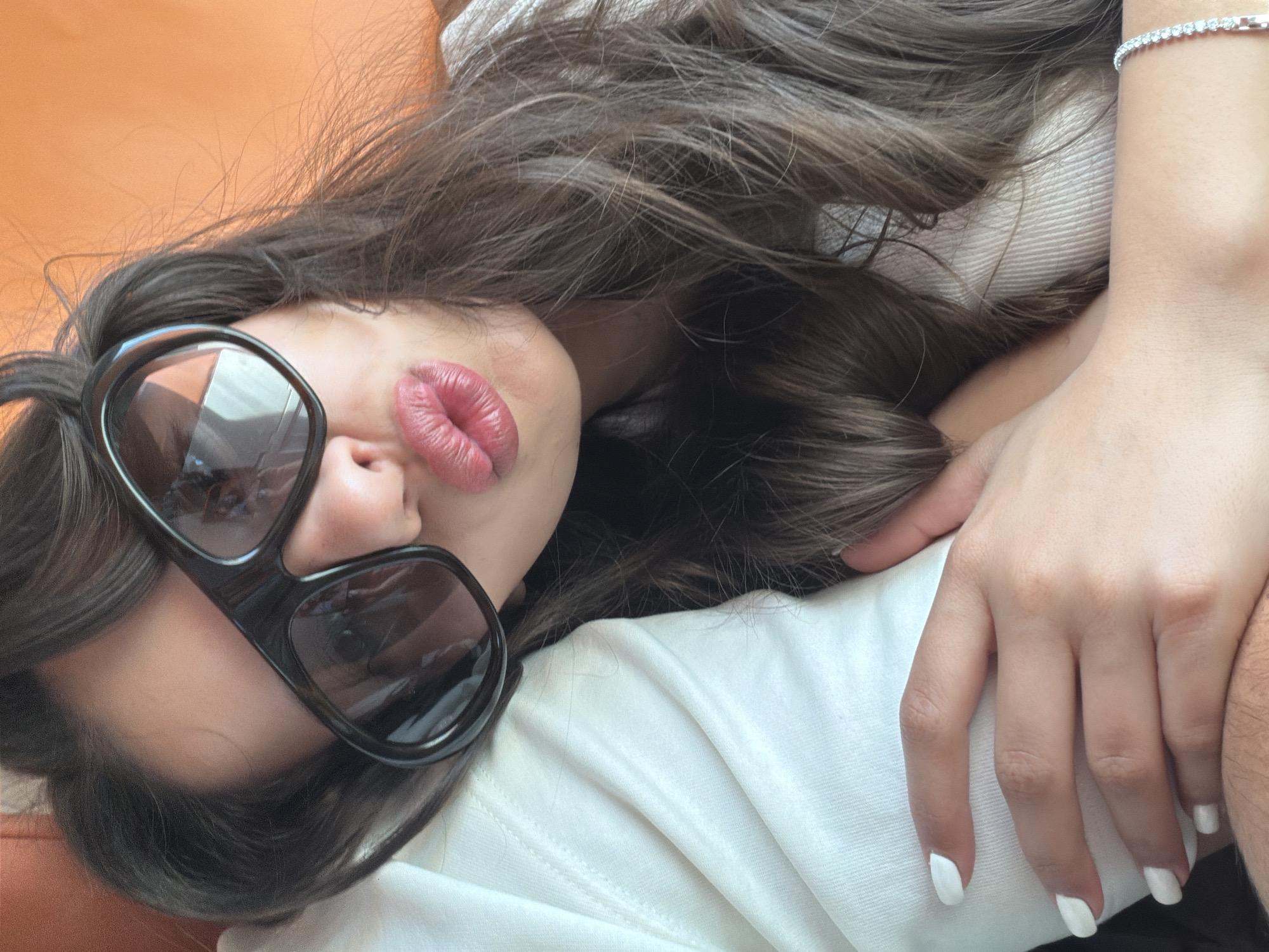 A close-up, high-angle shot captures a person, likely female, lying down in Cagnes-sur-Mer, France. Her dark, wavy hair cascades around her head, framing her face. She is wearing large, black-framed sunglasses, reflecting light and partially obscuring her eyes and upper face. Her lips are a prominent feature, colored with red or deep pink lipstick and puckered into a playful kiss pose.
She is dressed in a white garment, possibly a t-shirt or sweater, with visible fabric folds. A shimmering diamond-studded bracelet adorns her left wrist. Her left hand, with neatly painted white fingernails, rests on what appears to be another person's arm, indicated by the darker, slightly hairy skin visible at the bottom right of the frame. The background directly behind her head is a solid, warm orange color, suggesting a couch, pillow, or wall. The scene conveys a relaxed and casual moment, taken in what appears to be daytime, given the use of sunglasses. No text is visible in the image.