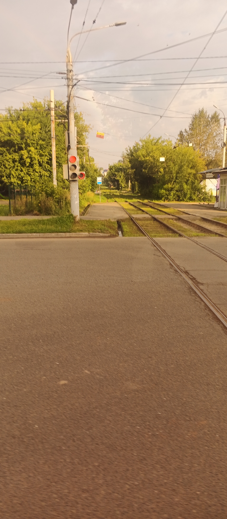The image depicts a street scene in Izhevsk, Russia, focusing on a tramway intersection. In the foreground, a paved road leads towards tram tracks that run parallel to a grassy verge. A traffic light for trams is visible on the left, with the red light illuminated, indicating a stop. Beyond the tram tracks, a sidewalk runs alongside a dense line of green trees. In the distance, a small building, possibly a tram stop shelter, with a purple and white facade, is situated to the right of the tracks. Overhead, a complex network of power lines crisscrosses the sky. The sky is overcast with soft clouds. The overall impression is one of a typical urban tramway crossing in a moderately vegetated area.