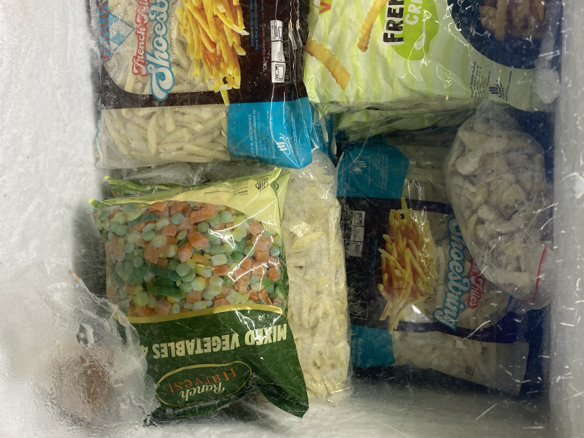 The image shows a close-up view of a freezer chest filled with various frozen food items. The food items are packaged in plastic bags and appear to be mostly fries and mixed vegetables. Some of the bags have visible labels, including one that says "Ranch Harvest Mixed Vegetables" and another that appears to be for "French Fries Shoestring." The freezer chest itself is covered in frost, and the lighting suggests an indoor environment, likely a commercial or domestic freezer. The image does not contain any information about the city of Payatieng, Indonesia.