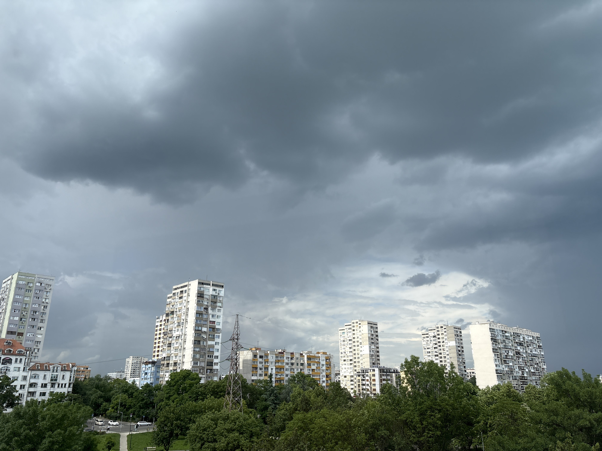 The image shows a cityscape under a stormy sky. The sky is dominated by dark gray storm clouds. Below the clouds are multiple white high-rise apartment buildings. The buildings are set behind a line of green trees. In front of the trees is a sidewalk and park. The buildings are located in Burgas, Bulgaria.