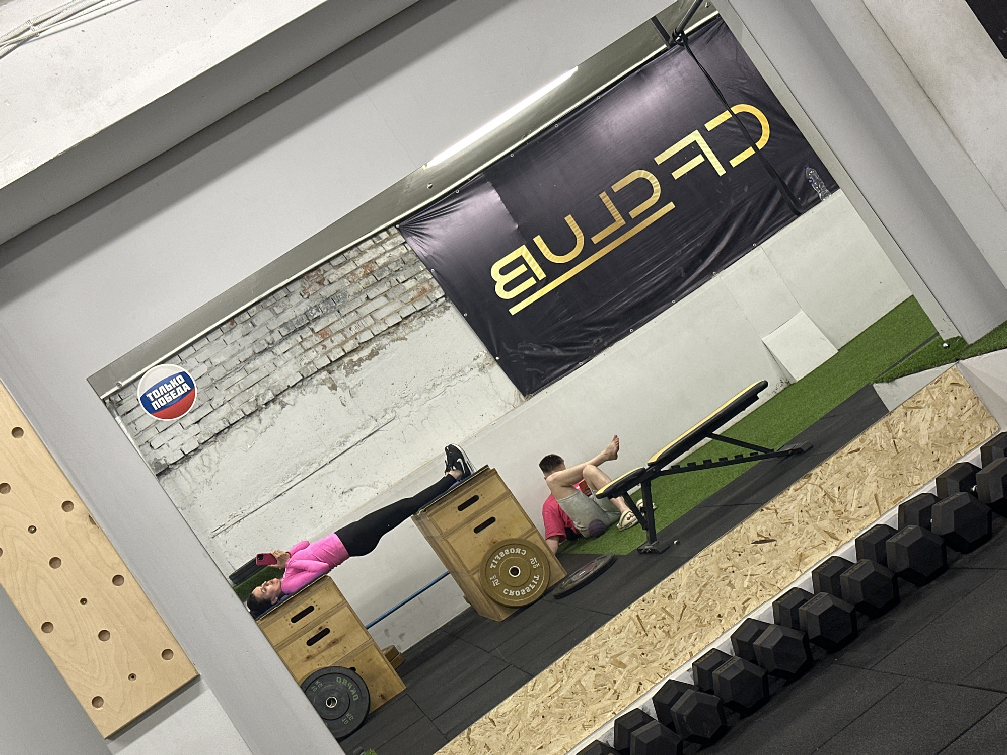 Gym interior in Chaikovsky, Russia. A woman in pink and black clothing is positioned on wooden boxes, holding a phone. A man in pink and grey clothing is on the floor near a weight bench. The wall has a banner that says "CF-CLUB". Dumbbells are arranged along the edge of a wooden structure. Another wooden structure is on the wall to the left. Weight plates are seen adjacent to the wooden boxes.