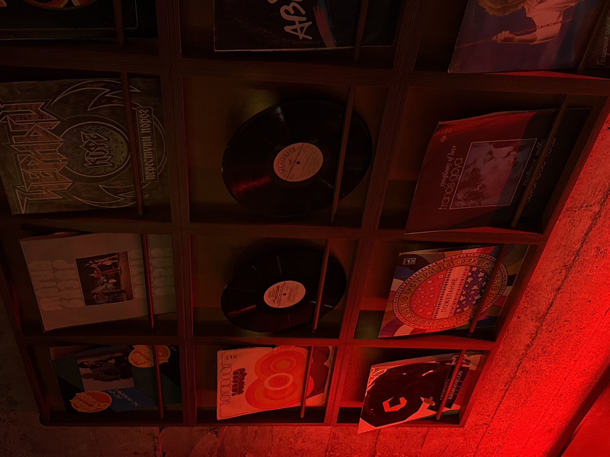 The image depicts a wooden shelving unit filled with vinyl records, likely in a store or private collection in Yerevan, Armenia. The records have a variety of album covers, including one in Cyrillic script that reads "Переступи Порог" (Perestupi Porog). Some records feature labels in Cyrillic, such as "Мелодия" (Melodiya), a Soviet record company. Other recognizable labels include "Francis Goya - Symphony of Love," "Armonicos", and AB. The records are arranged in individual compartments within the shelving unit. The scene is illuminated by red lighting, which creates a specific atmosphere. The focus is on showcasing the collection and variety of vinyl records stored in the unit.