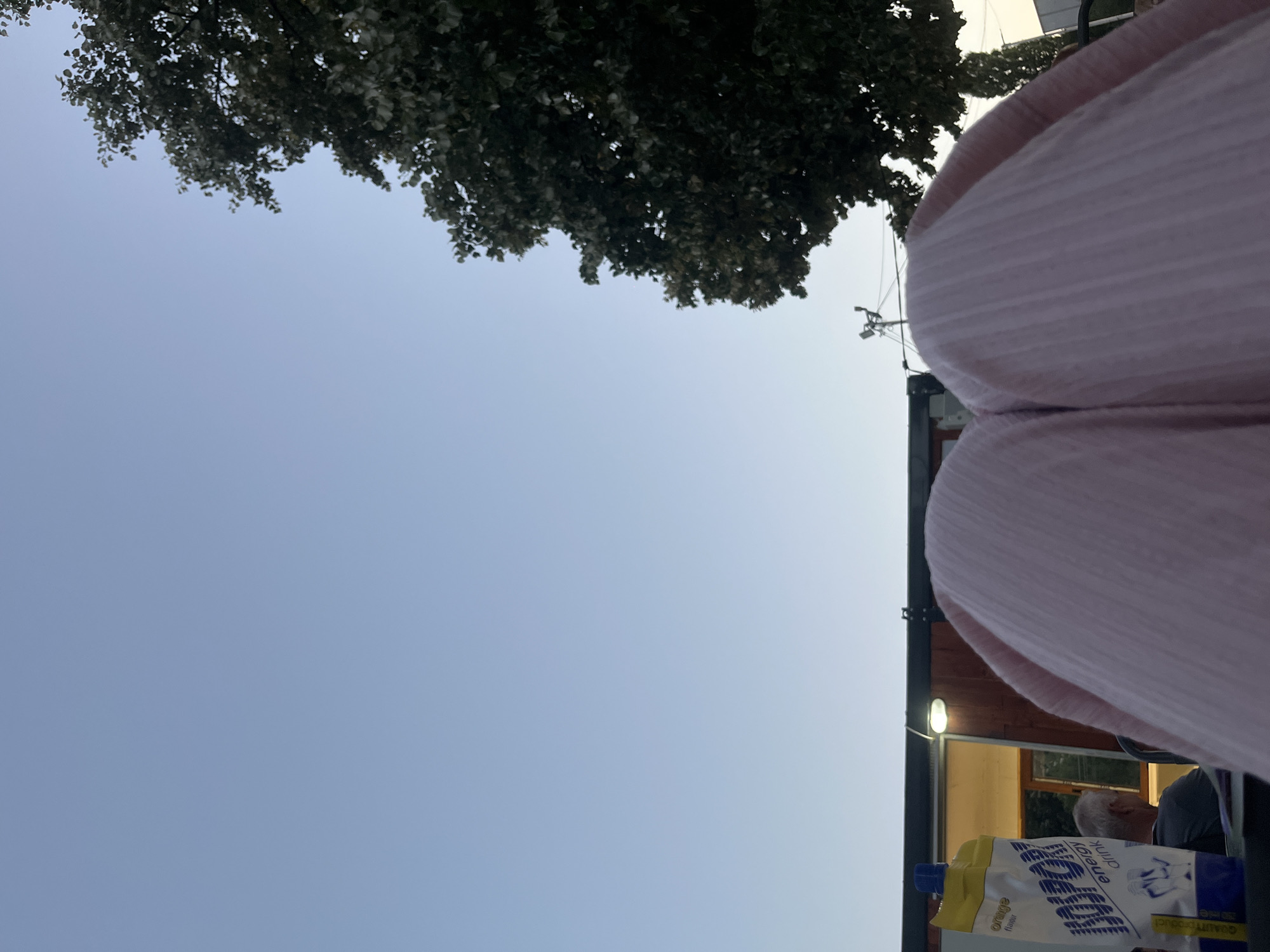 The image shows a close-up view of a person's knees, wearing pink ribbed trousers, positioned in the foreground. To the left, a white and yellow pouch of "ISOSPORT energy drink" with orange flavor is visible. Behind the drink, a person with gray hair is seen sitting, facing away from the camera. The background features a light-colored sky and the dark foliage of a tree on the right. A building with a window and a light fixture is visible behind the drink pouch. The scene appears to be outdoors, possibly during sunset, given the lighting. The specific location in the city of Vratsa, Bulgaria cannot be determined from the visual information provided.