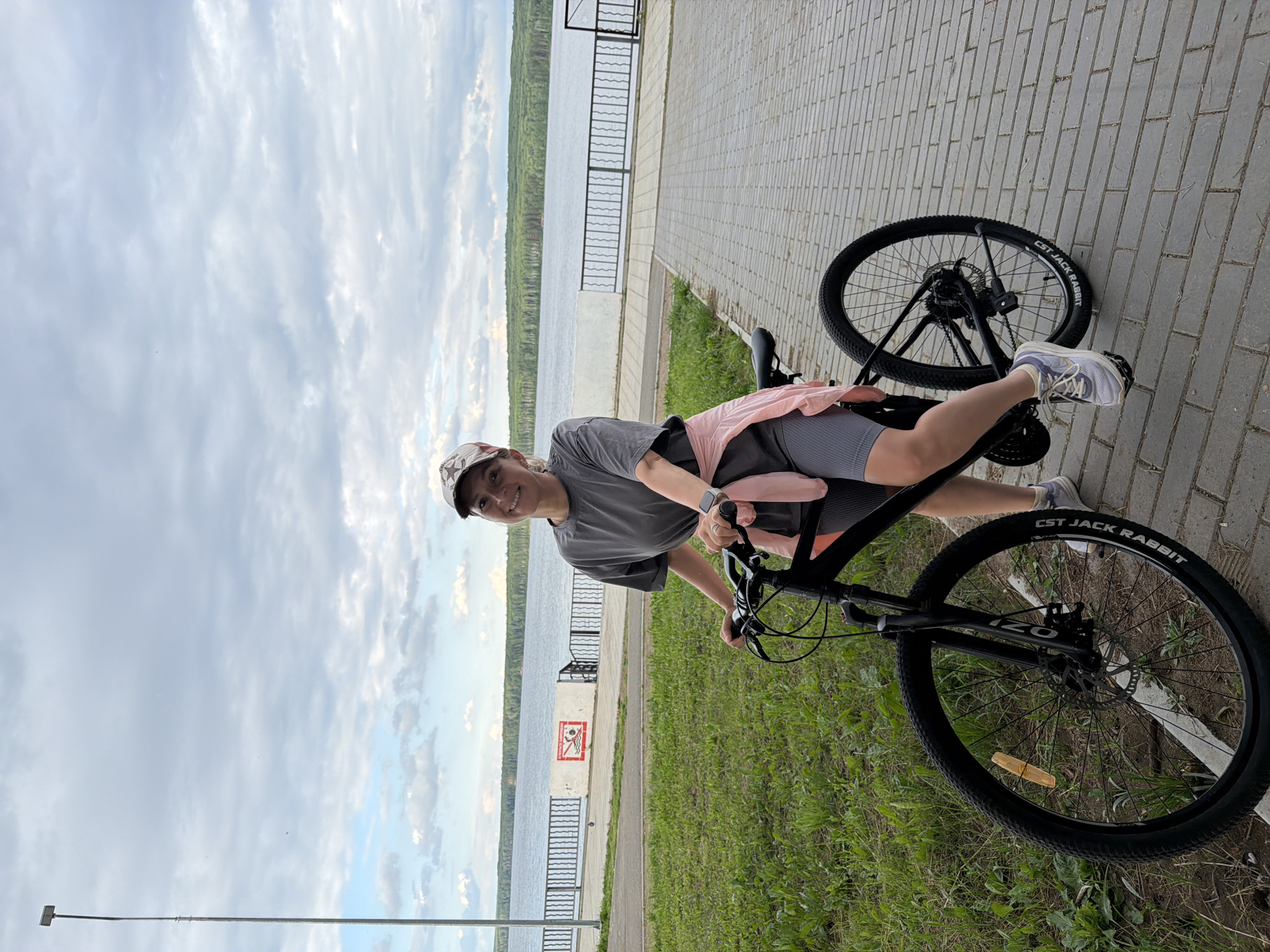 A woman in athletic attire is posing on a black bicycle on a paved walkway. She is wearing a grey t-shirt, grey shorts, a pink jacket tied around her waist, and a camouflage baseball cap. The bicycle has a prominent front wheel with a disc brake. In the background, a body of water stretches to the horizon, lined with trees. A metal fence runs along the edge of the walkway. The sky is cloudy. The location appears to be a recreational area, possibly near a waterfront, in the city of Chaikovsky, Russia.