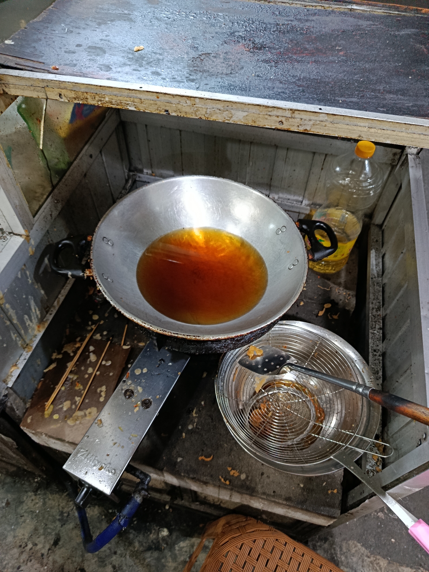 The image depicts a street food stall, likely in Depok, Indonesia, specializing in deep-fried food. A large, well-used aluminum wok filled with bubbling hot oil dominates the center of the frame. Next to it, a stainless steel sieve holds fried food, with a slotted spoon resting on top, suggesting food is being drained or prepared for serving. In the background, a large plastic bottle of cooking oil is visible. The stall itself appears rustic, with wooden and metal components, and there are remnants of food debris and oil splatters on the surfaces, indicative of an active food preparation area. The overall scene conveys a sense of everyday commerce and the preparation of popular street food.