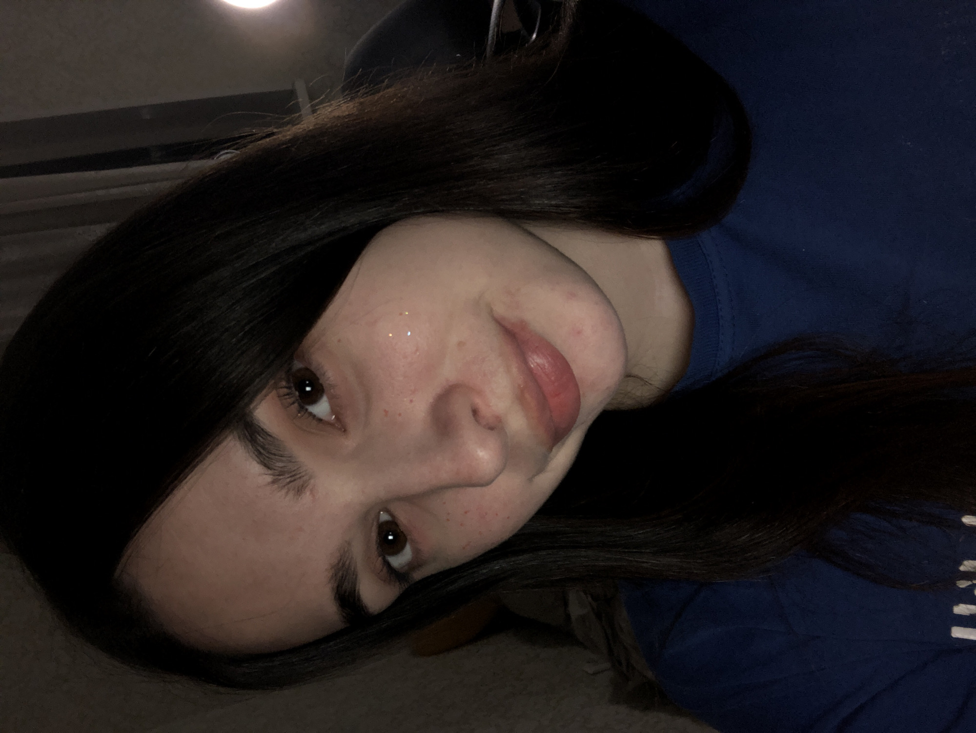 A young woman with dark hair and brown eyes is looking upwards. She has fair skin with freckles. She is wearing a dark blue t-shirt. The background is indistinct, but suggests an indoor setting with a light source. There is no evidence to suggest the location is Noviy, Russia.