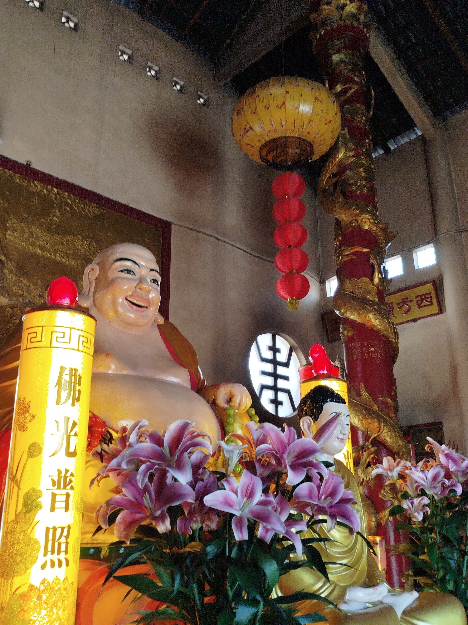 The image depicts a scene inside a Buddhist temple, likely in Da Lat, Vietnam, given the architecture and iconography. The main subjects are two Buddha statues: a large, smiling Buddha (likely a depiction of Budai, or the "Laughing Buddha") and a smaller Buddha statue, both surrounded by pink lilies. The smiling Buddha is positioned to the left of the frame and is made of a fleshy-toned material. Next to the Buddha is a yellow column-like object, which has Chinese characters, in gold, displayed on it. The smaller Buddha statue is golden and sits in a meditative pose.
In the background, there is a red and gold pillar with a dragon design, a yellow lantern with red pendants hanging from it, and an ornate screen. A sign with Chinese characters is fixed to the wall. The overall environment is dimly lit, and the colors are rich, with gold, red, and yellow dominating. The composition suggests a place of worship or veneration. The building’s ceiling is a mixture of wooden beams and slats and a concrete ceiling with small windows. The roof structure seems to be old and in need of repair.
