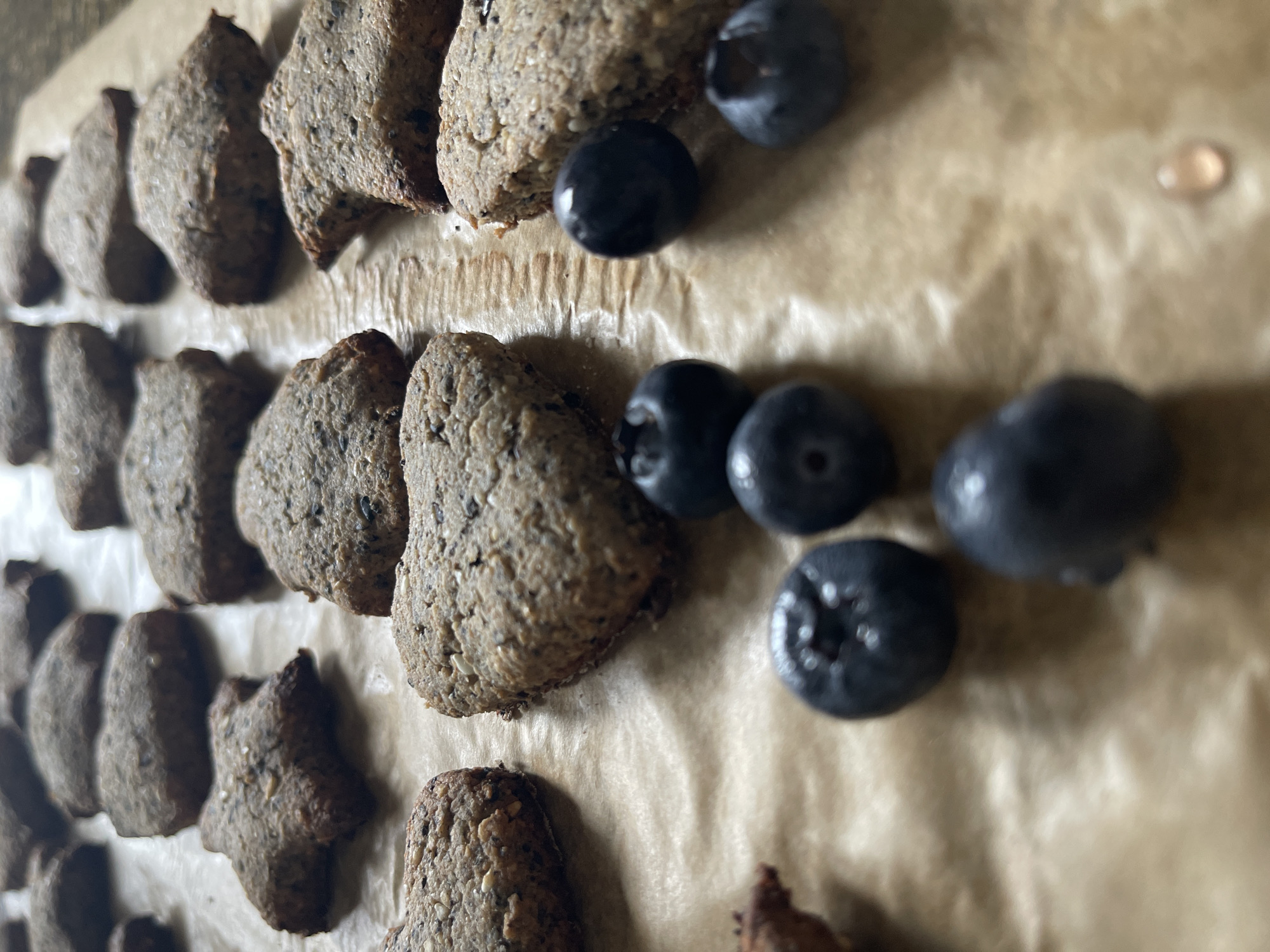 The image shows a close-up of several dark-colored cookies and blueberries arranged on a sheet of parchment paper. The cookies are irregularly shaped, with some resembling triangles and others appearing to be abstract shapes like stars. They appear to be textured and slightly rough. Scattered around the cookies are a few fresh blueberries. The parchment paper is light brown and crinkled, with a single drop of liquid visible in the upper right corner. The scene is indoors and appears to be naturally lit. No people are visible.