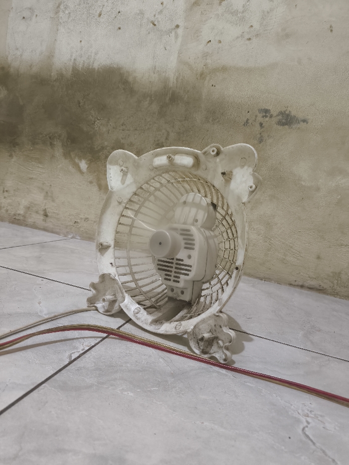 A dirty, white plastic desk fan sits on a tiled floor against a mottled wall. The fan lacks its front grill, exposing the blades and motor. A power cord with red and yellow wires is visible in the foreground. Dust and grime accumulate on the fan's surface, particularly around the base and grill supports, giving it a neglected appearance. The floor tiles are light gray and appear worn.