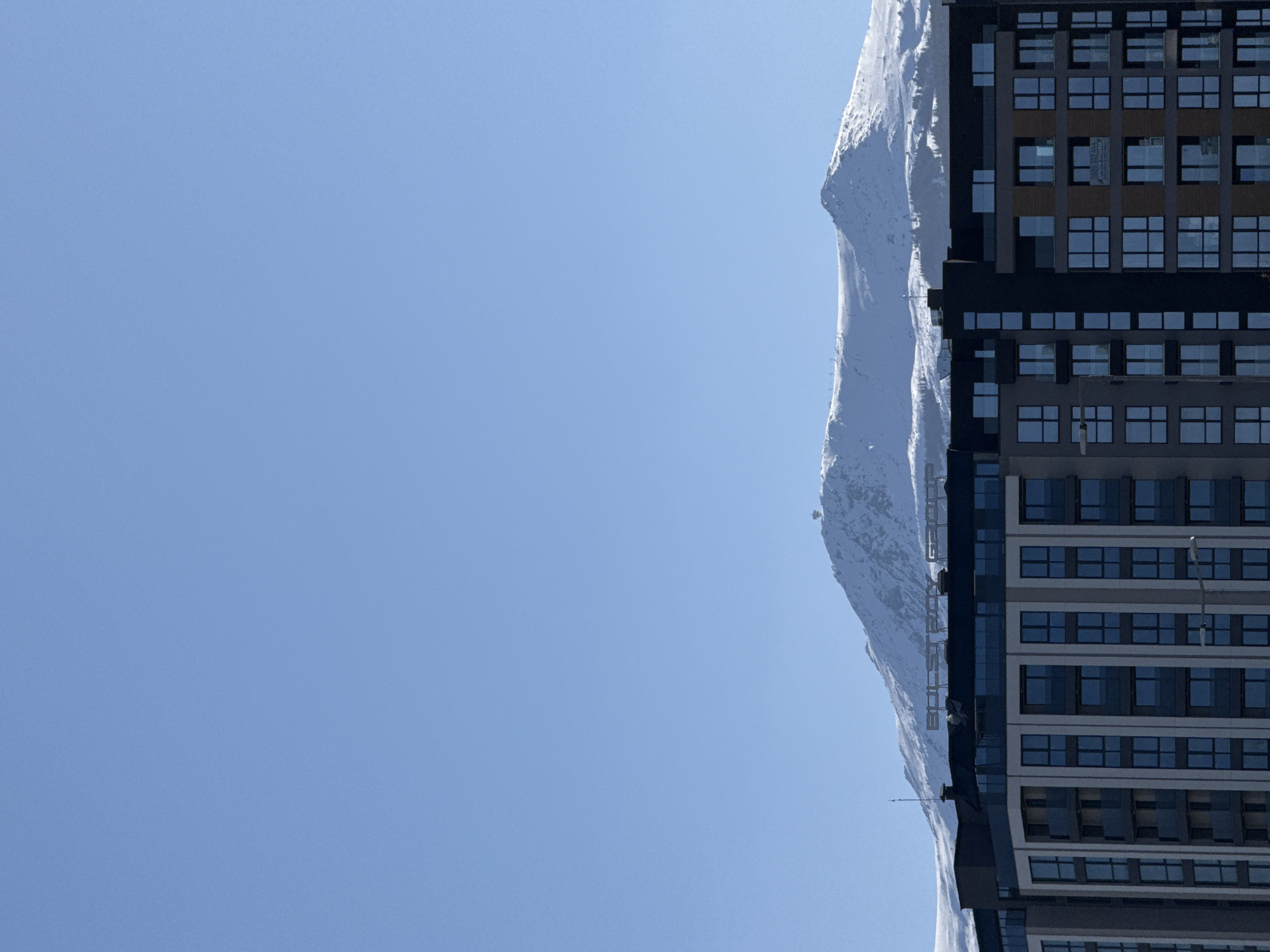 A low-angle shot depicts a building with numerous windows next to a snow-covered mountain. The building is a modern structure, featuring a uniform grid of rectangular windows across multiple floors. The exterior facade is a combination of dark and light neutral tones. Adjacent to the building is a mountain covered in snow. The sky is clear and blue, taking up a significant portion of the frame. The image is taken outdoors on a clear day with ample sunlight. The photo location is not Sofia, Bulgaria.