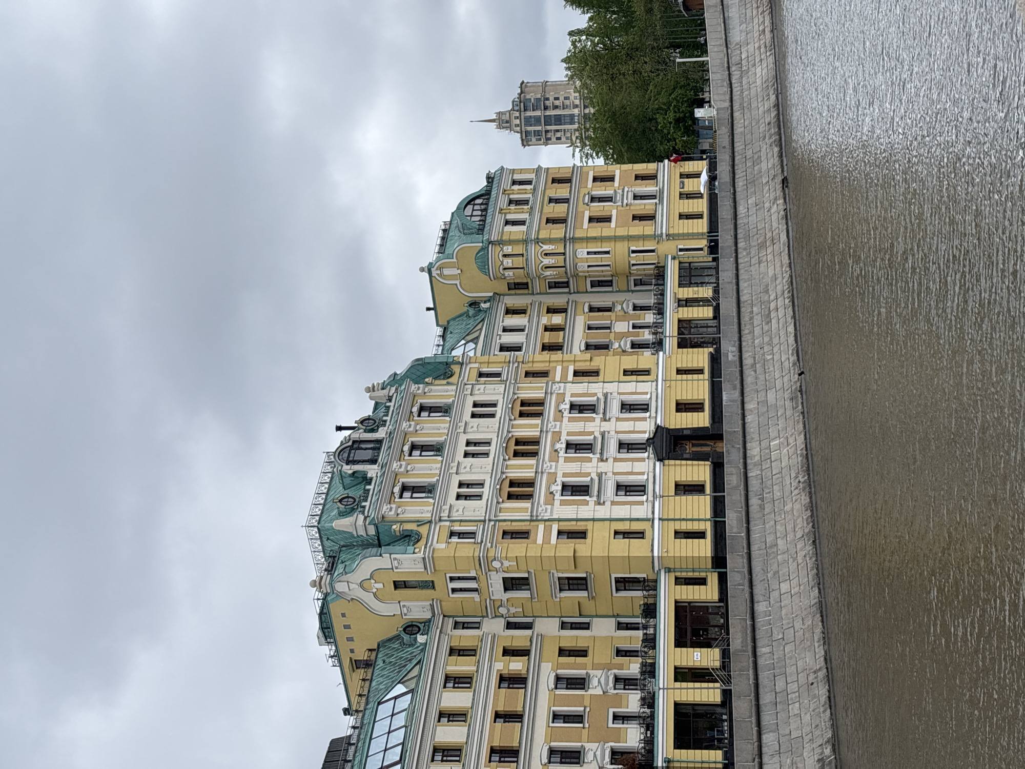 The image depicts a building complex on the embankment of the Moskva River in Moscow, Russia. The buildings are multi-story, with a yellow facade and distinct architectural details, including arched windows, decorative moldings, and green roofs. The sky is overcast, with a gray, cloudy sky overhead. The river is visible on the right side of the frame, its surface reflecting the cloudy sky. A concrete embankment separates the building from the water. Another building can be seen in the distance behind the trees.