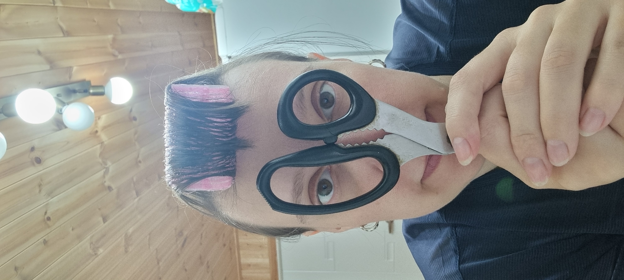A woman is posing for the camera, holding a pair of scissors in front of her eyes. She is wearing hair rollers. The background includes a wooden wall with lights and a white door. The location cannot be determined to be Dunnae-myeon, South Korea from the image.