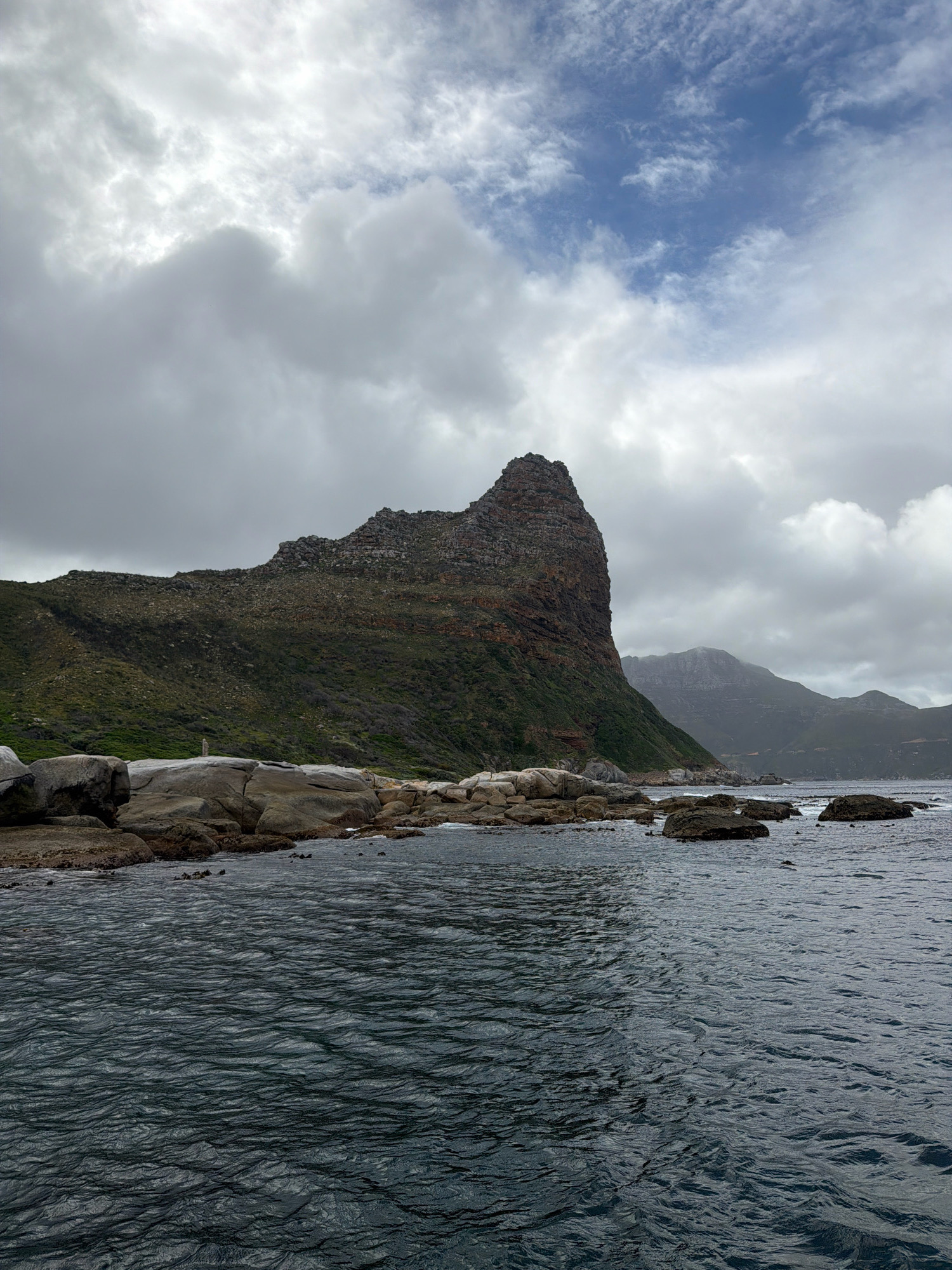 The image depicts a dramatic coastal landscape in Hout Bay, South Africa, likely near Chapman's Peak. In the foreground, the dark, choppy water of the Atlantic Ocean stretches across the frame, its surface rippling under an overcast sky. The shoreline is characterized by large, weathered boulders and smaller rocks, some submerged by the tide.
Dominating the midground is a steep, rugged mountain formation, covered in dense, green vegetation. The peak is sharply defined, its rocky texture contrasting with the softer slopes. To the right and further in the distance, more mountainous terrain rises, shrouded in a hazy, muted light.
The sky above is a mixture of heavy, grey clouds with patches of brighter sky visible through them, suggesting a day with variable weather. The overall impression is one of raw, natural beauty and the powerful presence of the South African coastline. There are no people or discernible man-made structures visible in the scene.