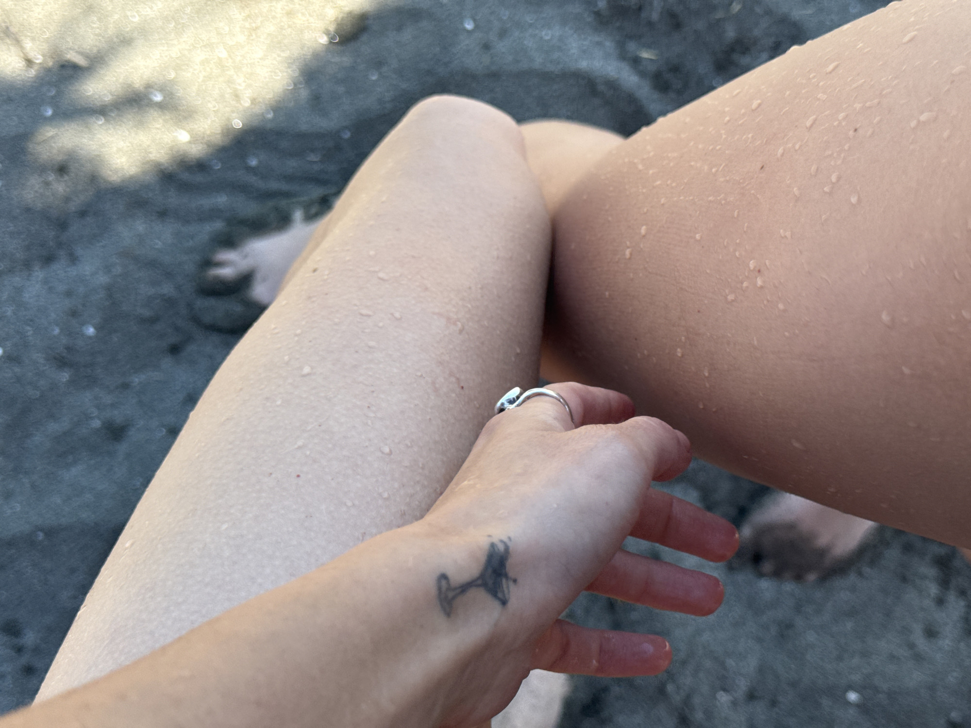 The image shows a close-up of a person's legs and hand. The skin on the legs is wet, with many small water droplets visible. A tattoo of what appears to be a wine glass is visible on the back of the hand. A silver ring with a blue stone is worn on one of the fingers. In the background, out of focus, is dark, wet sand, and a faint outline of a foot is visible. The lighting suggests it is daytime, with some dappled sunlight. The location of Burgas, Bulgaria is not discernible from the image itself.