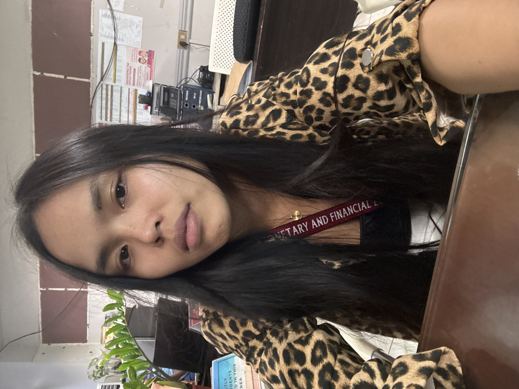 A young woman with long, dark hair is taking a selfie indoors. She is wearing a leopard-print jacket over a dark top, and a lanyard with text is visible around her neck. She is sitting at a desk in an office environment, with a plant and office equipment visible in the background. The wall behind her appears to be decorated with papers and a poster. There is a desk in front of her. The setting appears to be an office in Davao City, Philippines.