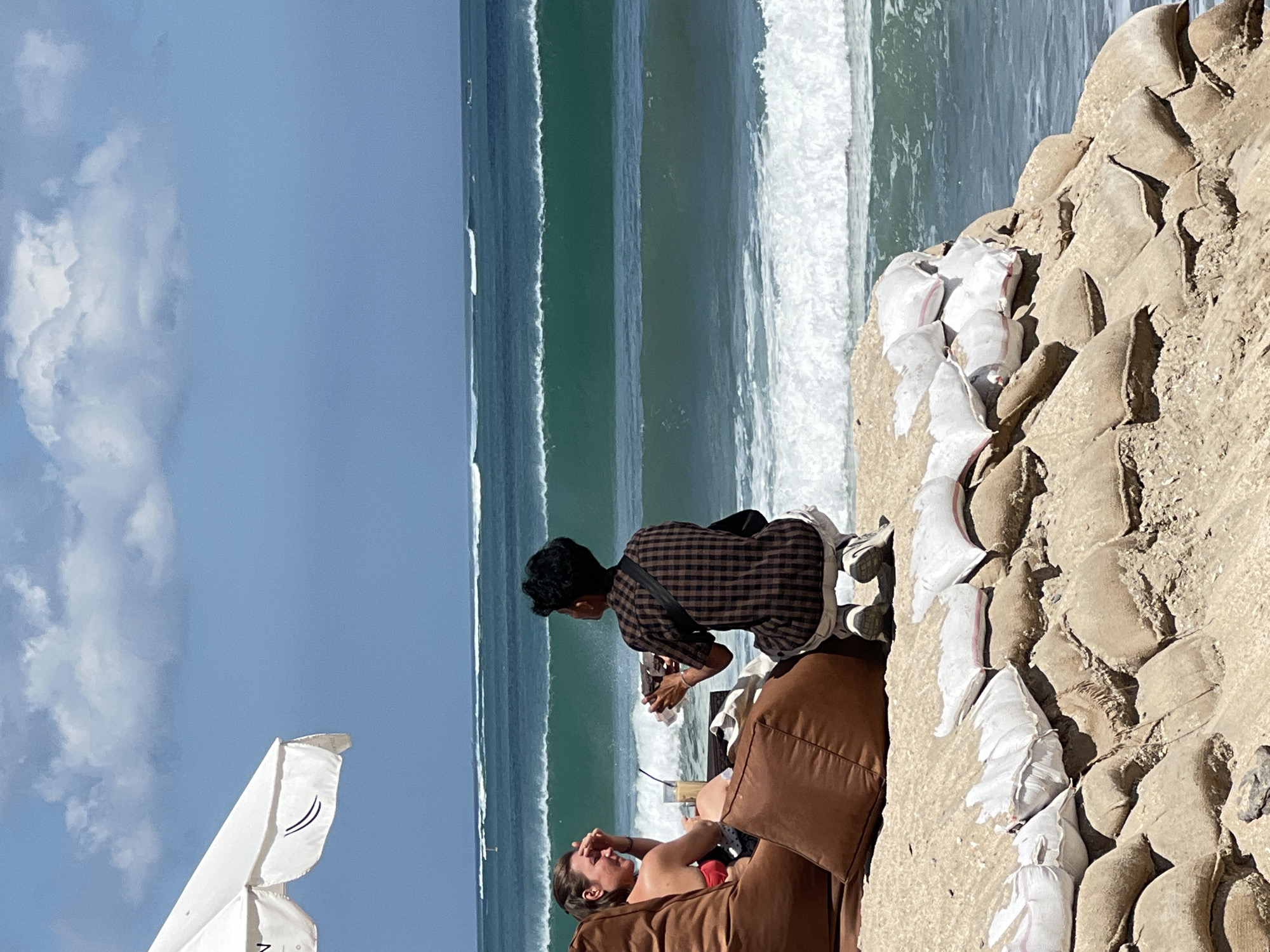 This image depicts a beach scene in Kuta, Indonesia, characterized by a protective sandbag barrier along the shore. In the foreground, stacked sandbags, some covered with white fabric, form a sturdy embankment against the ocean. Beyond the barrier, two individuals are seated on a large, brown beanbag chair on the sandy beach, facing the ocean. A woman in a red bikini is on the left, gesturing with her hand, while a person in a brown checkered shirt is to her right, holding a drink and a phone. In the background, the turquoise ocean displays multiple lines of breaking waves under a bright, partly cloudy sky. A white beach umbrella is visible on the far left, providing shade. The overall environment suggests a relaxed, tropical beach setting with measures in place to mitigate coastal erosion.