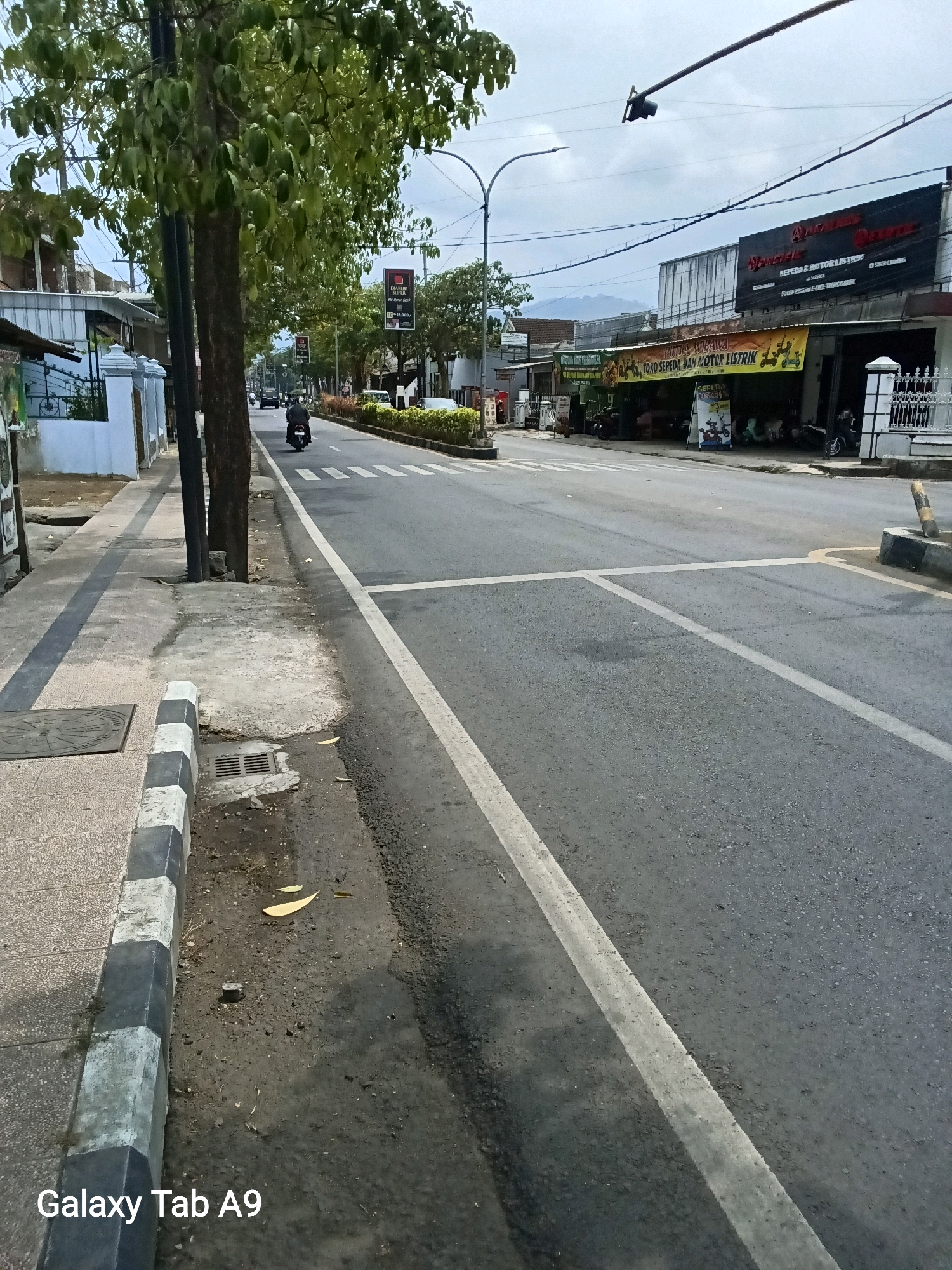 A view of a street in Kelutan, Indonesia, showing a paved road with clear lane markings, a sidewalk on the left, and buildings and trees lining the street. A single motorcycle rider is visible on the road, approaching a crosswalk. The sky is overcast, and the overall lighting suggests it is daytime. Several businesses are identifiable, including "Pacific" which appears to be a motorcycle dealership and service center, with signage in Indonesian. The sidewalk has a textured surface and a concrete curb with alternating black and white painted sections. A storm drain is present near the curb. The scene captures a typical urban street environment with elements of local commerce and daily life.