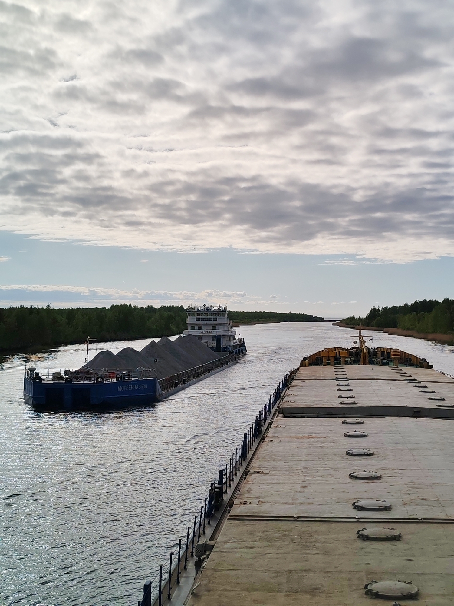 The image shows a river scene in the city of Ozernoe Ust'e, Russia. Two large barges are on the water, with the closest one taking up most of the frame. This barge has a gray, concrete-like surface with evenly spaced round hatches. A dark metal railing runs along its edge. The second barge, further away, is loaded with large mounds of gray material, possibly gravel or ore. It is being pushed by a tugboat. The river is bordered by green trees on both sides. The sky is overcast with gray and white clouds.