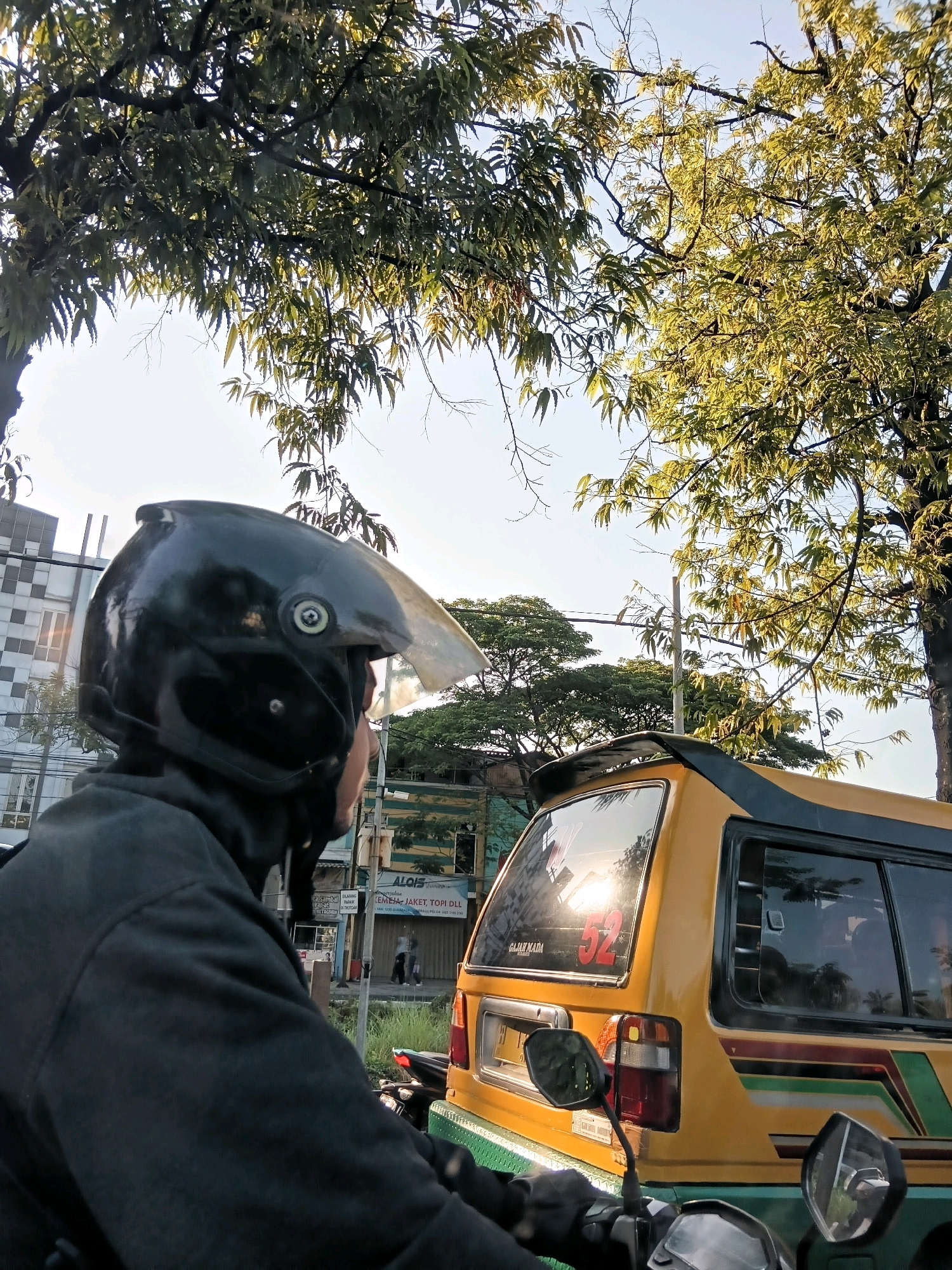 The image shows a perspective from a person riding a motorcycle, looking towards the back of a yellow van in Surabaya, Indonesia. The rider is wearing a black helmet and a dark jacket. The yellow van has a number "52" visible on its rear window, which is reflecting the sunlight. The scene is outdoors, with trees overhead and buildings in the background. The overall lighting suggests it is daytime, likely late afternoon due to the warm sunlight. The environment appears to be an urban street.