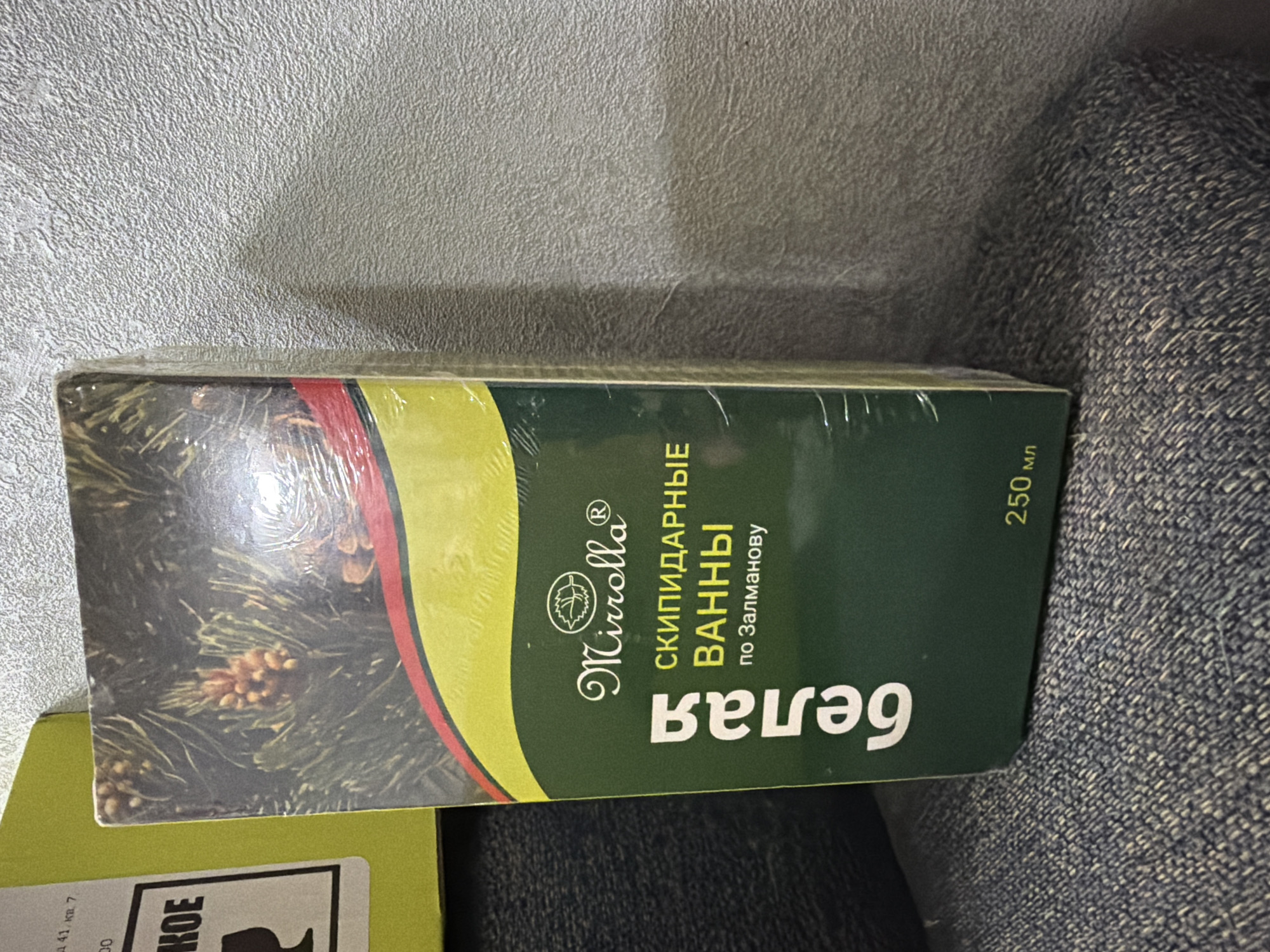 This media file shows the front of a boxed product, likely a bath product due to the text on the packaging. The box is green with a yellow and red striped design on the left side. A portion of a pine branch with cones is visible on the far left of the box, suggesting a natural or herbal theme.
The text on the box is in Russian and reads:
"MirrollaⓇ"
"СКИПИДАРНЫЕ ВАННЫ" (Turpentine Baths)
"по Залманову" (According to Zalmanov)
"белая" (White)
"250 мл" (250 ml)
The product is wrapped in clear plastic. The box is lying on a textured surface, possibly a wall, and next to a blue, textured fabric, possibly denim or a similar material. A portion of another yellow-boxed item is visible at the bottom left, with some Russian text that is partially obscured, including "141, кв. 7", "00", and "КОЕ".
The lighting suggests an indoor setting, with no specific time of day or weather information inferable. The focus is on the product packaging, with no people or animals visible. The overall impression is that of a product advertisement or a photograph of a purchased item.