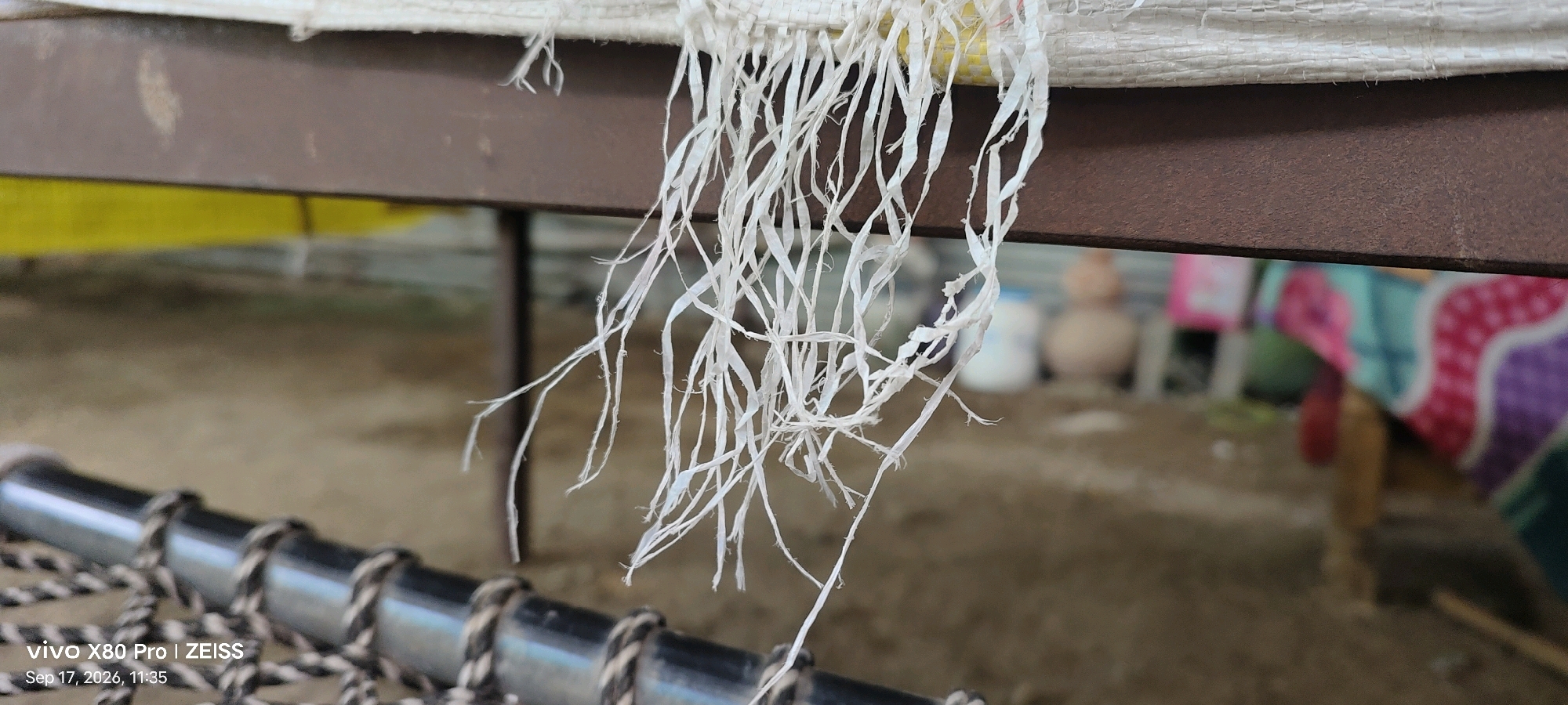 The image is a close-up shot of frayed white plastic strings hanging from the edge of a white woven material, possibly a bag or mat. These strings are delicate and appear to be unraveling. They are suspended above a brown metal bar, which is part of a structure that includes a woven black and white cord. In the background, the scene is out of focus, showing a ground surface and colorful objects, some of which appear to be fabrics or blankets, and possibly some spherical items. The bottom left corner of the image displays a watermark indicating "vivo X80 Pro | ZEISS" and the date "Sep 17, 2026, 11:35". The location is identified as Guradiya Mana, India.