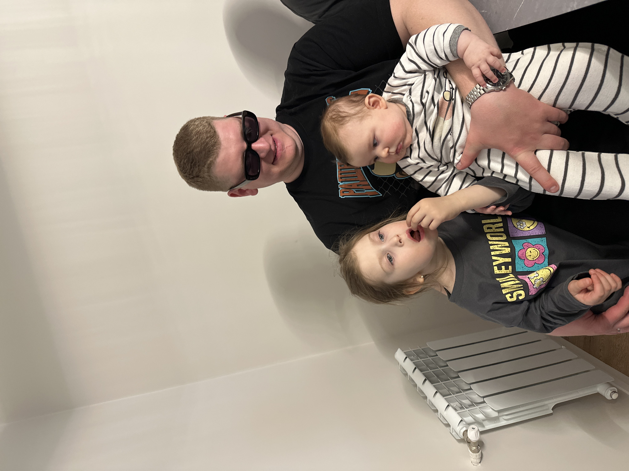 The image captures an indoor family scene featuring a man, a baby, and a young child, likely in a home in Chaikovsky, Russia. The photograph is oriented vertically but depicts the subjects as if rotated 90 degrees counter-clockwise against a plain, light-colored wall.
At the center, a man with short, light brown hair and black sunglasses looks towards the camera with a subtle smile. He wears a black t-shirt with a partially visible graphic on the chest and a silver watch on his left wrist. He is holding a baby dressed in a white and black striped long-sleeved onesie, who appears calm and is looking downwards or to the side.
Positioned below the baby, a young child with light brown hair and blue eyes is visible, looking slightly upwards with an open-mouthed expression, possibly in surprise or mid-speech. This child is wearing a gray long-sleeved t-shirt that clearly displays the text "SMILEY WORLD" in yellow lettering, complemented by colorful flower and smiley face graphics.
In the foreground, towards the bottom right of the image, a modern white radiator with horizontal slats is visible on the floor, connected by a pipe and valve. The lighting is bright and even, suggesting a daytime setting indoors. The plain walls provide a simple backdrop, emphasizing the family members.