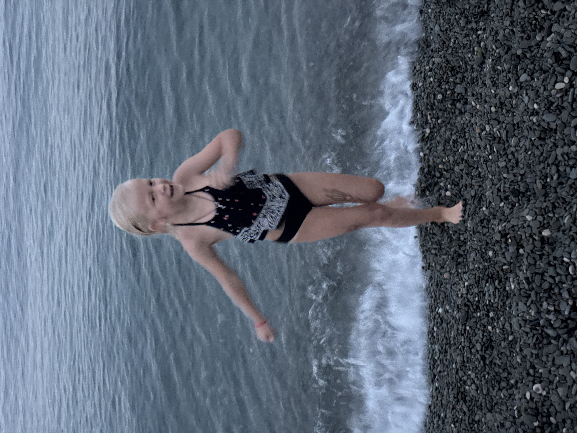 A young girl with blonde hair is pictured from a high angle, partially submerged in the ocean. She is wearing a black and white patterned swimsuit and appears to be mid-stride, with one leg extended and the other bent. Her mouth is open, and she is looking towards the camera with a joyful expression. The water is calm and reflects the overcast sky, creating a textured blue surface. Along the right side of the image, a dark, pebbled beach meets the water. The pebbles are small and rounded, varying in shades of gray and black. White foam from a gentle wave laps at the edge of the shore and the girl's legs. The lighting suggests it may be a cloudy day. A thin pink bracelet is visible on the girl's left wrist. There is a tattoo on her right thigh.