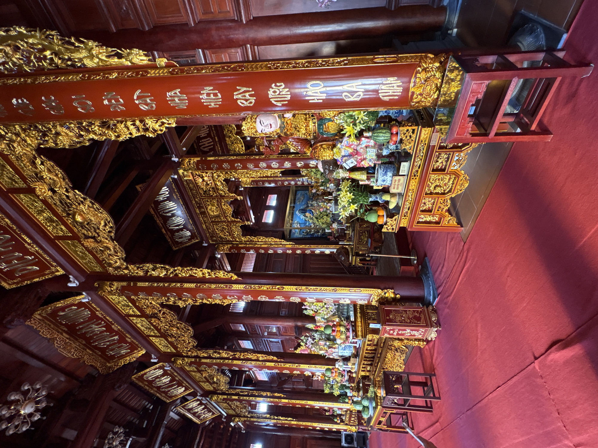 The image captures the interior of a building in Nam Nha Trang Ward, Vietnam. The decor is elaborately ornate, with a dominant color scheme of red and gold. Red pillars and beams are adorned with gold lettering and intricate gold carvings.
Several levels of structures rise towards the ceiling, each decorated with similar red and gold elements. Fruits, flowers, and other offerings are arranged on these levels, some near statues.
A red carpet covers the floor, partially obscuring the base of some of the structures and a small wooden staircase. The overall impression is one of reverence and tradition.