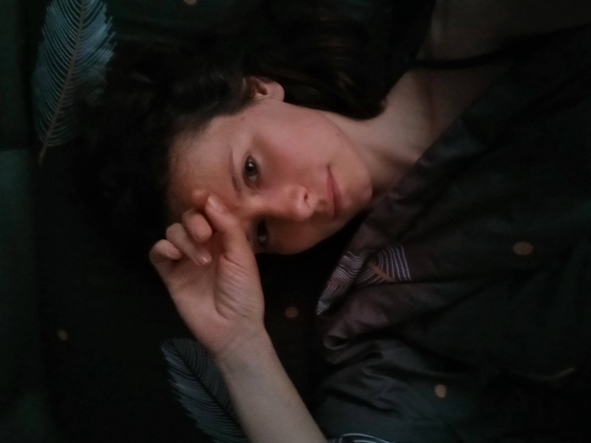 A young woman with dark hair lies on her back, her right hand resting on her forehead. She is partially covered by a dark duvet with a feather pattern. The scene is dimly lit, with the background being a dark, out-of-focus expanse. The woman is looking directly at the camera. The image appears to have been taken indoors, in a domestic setting. There is no discernible information that allows for the specific location in the city of Yekaterinburg, Russia to be identified.