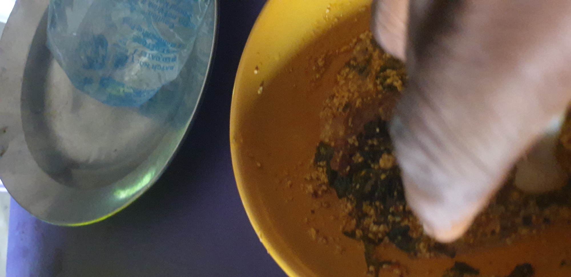 An overhead shot captures a rustic, rustic dish served in a yellow bowl on a dark surface, likely a table. The dish appears to be a coarse grain with visible green leafy vegetables and possibly some red seasoning, suggesting a savory, possibly African, cuisine. In the upper background, a metal plate sits on the same dark surface, holding a clear plastic bag filled with a blue liquid, possibly a beverage or ice pack. A person's hand, with its skin tone and texture, is partially visible in the foreground, reaching into the yellow bowl, suggesting the act of eating or preparing to eat the dish. The lighting is dim and uneven, creating a slightly grainy texture throughout the image, indicative of a candid shot rather than a professional one. The overall setting seems informal, perhaps a casual meal at home or an outdoor eatery.