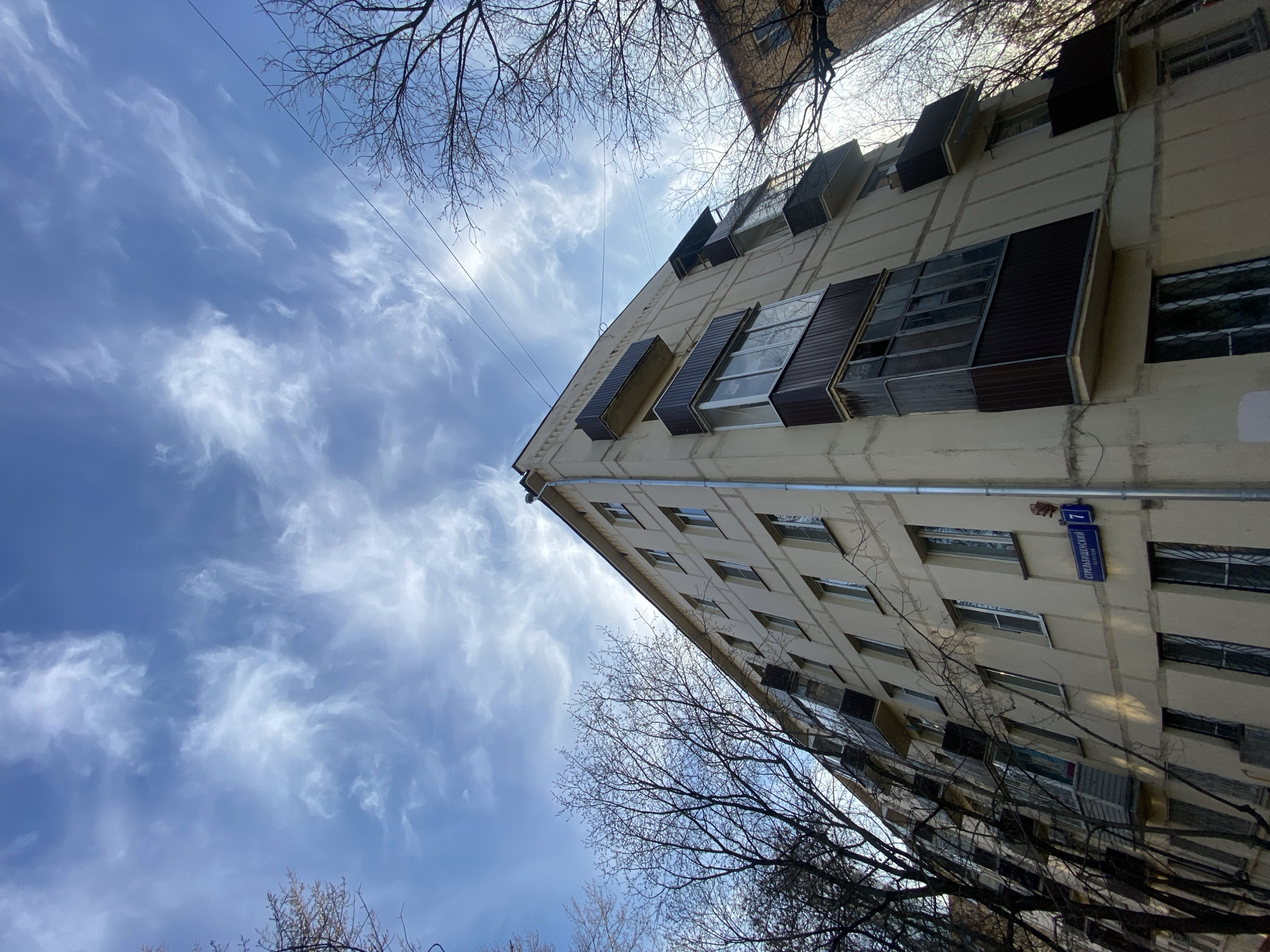 A low-angle exterior photograph captures a multi-story residential building at Strelbishchenskiy Pereulok 7 in Moscow, Russia, against a bright blue sky with wispy white clouds. The building, constructed of pale yellow/beige concrete panels, features at least five visible floors with rectangular white-framed windows. Several balconies are present; some are open concrete ledges, while others are enclosed and glazed with dark brown paneling. A prominent enclosed balcony spans two stories on the left side of the primary facade. A light-colored drainpipe runs down the building's right corner. A blue street sign on the facade identifies the location as "СТРЕЛЬБИЩЕНСКИЙ ПЕРЕУЛОК" (Strelbishchenskiy Lane) and indicates the number "7". Bare deciduous tree branches frame the building on both sides, and thin power lines traverse the upper right sky. No individuals or specific actions are visible in the scene.