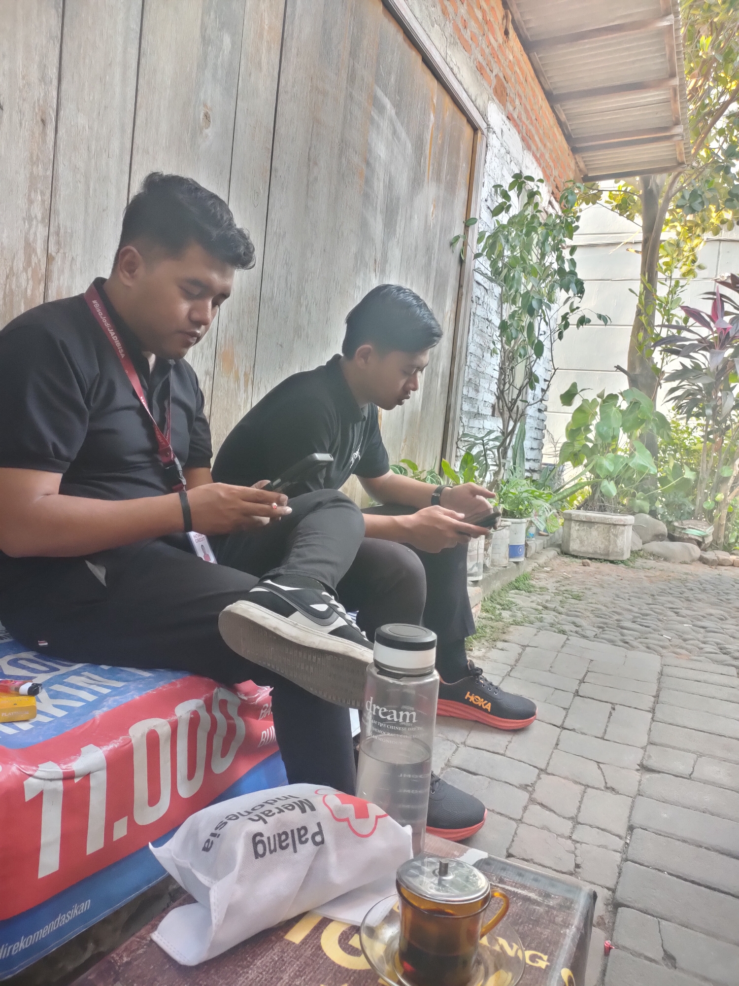 Two individuals, appearing to be adult males, are seated outdoors on a low, blue and red patterned surface. The person on the left, wearing a black polo shirt and black pants, is looking down at a mobile phone in their hands and appears to be holding a cigarette. A red lanyard with white text is visible around their neck. The person on the right, also in a black polo shirt and black pants, is also looking down at a mobile phone. Both are wearing athletic shoes; the person on the left has on black and white sneakers, while the person on the right is wearing black shoes with orange accents and the brand "HOKA" visible.
In front of them, on a weathered wooden surface, is a clear plastic water bottle with the label "dream" and text in a foreign language. Next to it is a small glass teacup with a metal lid containing amber liquid, placed on a saucer. A white, bagged item with red lettering and a cross symbol is also present, as is a yellow pack of cigarettes on the blue and red surface.
The background consists of a wooden wall, a brick wall with peeling white paint, and various potted plants, some with large green leaves and others with reddish-purple foliage. A tree with sparse green leaves is also visible. The ground is paved with grey cobblestones. The lighting suggests it is daytime, with sunlight casting shadows and illuminating parts of the scene. The overall impression is a casual outdoor setting, possibly a cafe or a communal area in Sooko, Indonesia, given the context.