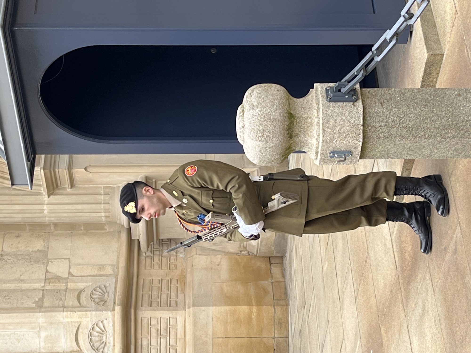 A soldier in a khaki green uniform stands guard outside a stone building. The soldier is wearing a black beret with a yellow emblem, polished black boots, and white gloves. He is holding a rifle. A stone bollard with a metal chain is to his right. An arched, dark blue doorway is behind the bollard and part of the building. The floor is composed of light-colored stone tiles. The architecture and presence of the guard suggest that this may be a governmental or important cultural site in the city of Luxembourg, Luxembourg.