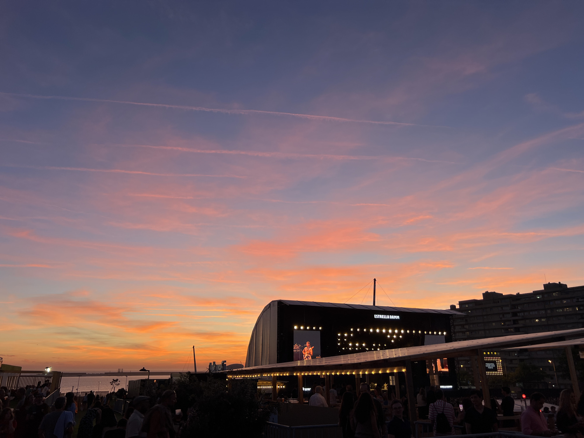 The image shows an outdoor concert venue at sunset in Porto, Portugal. The sky is a vibrant mix of orange, pink, and purple hues, with thin contrails streaking across it. In the background, a large stage is set up with a screen displaying a musician performing. The stage is illuminated by a line of lights. Below the stage, there are covered seating areas and a crowd of people gathered, many of whom appear to be watching the performance. In the far background, several multi-story buildings are visible, characteristic of an urban environment. The scene suggests a lively atmosphere at a public event.
