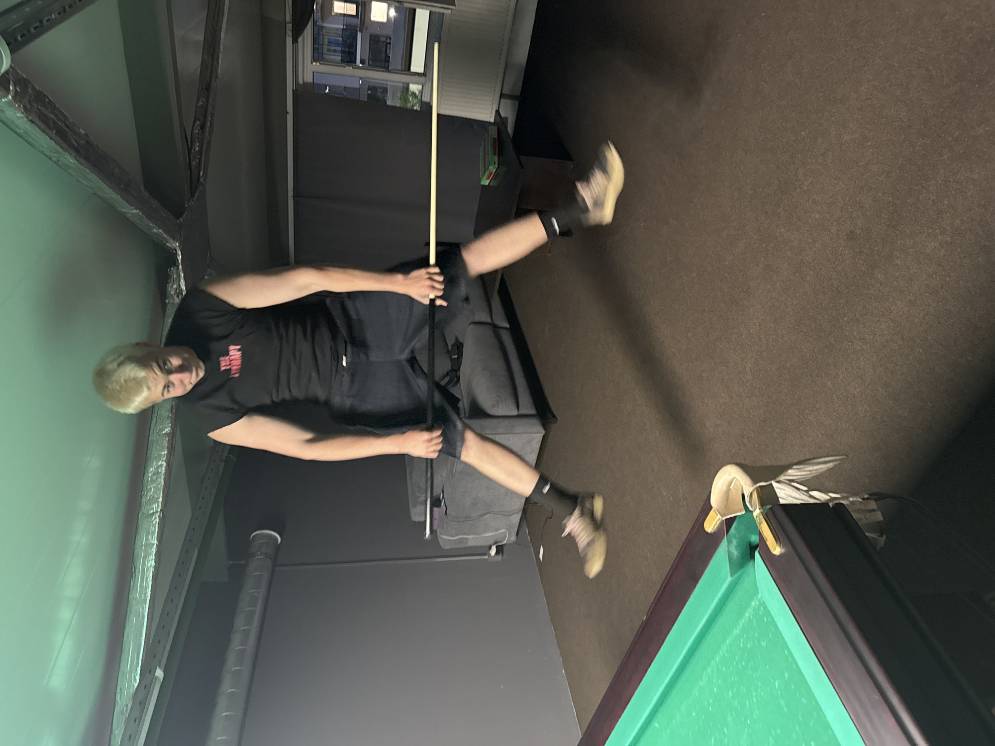An individual with blonde hair, wearing a black t-shirt and dark shorts, jumps in the air while holding a pool cue. The person is positioned in the center of the frame, with their legs extended as they jump. A green pool table is visible in the foreground to the left. In the background, a gray sofa and a black table can be seen. The environment appears to be an indoor recreational space, possibly a game room or basement, with exposed ceiling beams and ductwork. The lighting is dim.