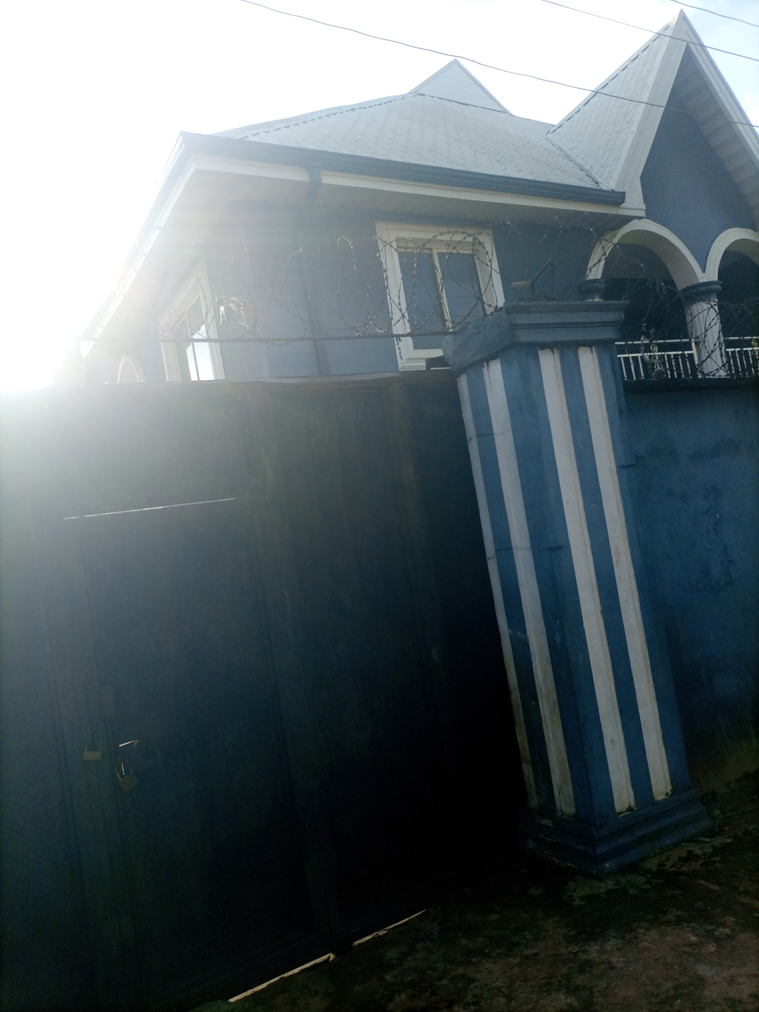 The image shows a blue house with white trim, located in Nkpor, Nigeria. The house has a multi-tiered roof with a prominent gable. There is a large metal gate in front of the house, and a decorative pillar with blue and white stripes stands to the right of the gate. Barbed wire is visible along the top of the wall. The sun is shining brightly, causing a lens flare on the left side of the image. The ground in front of the gate is paved with dark material.