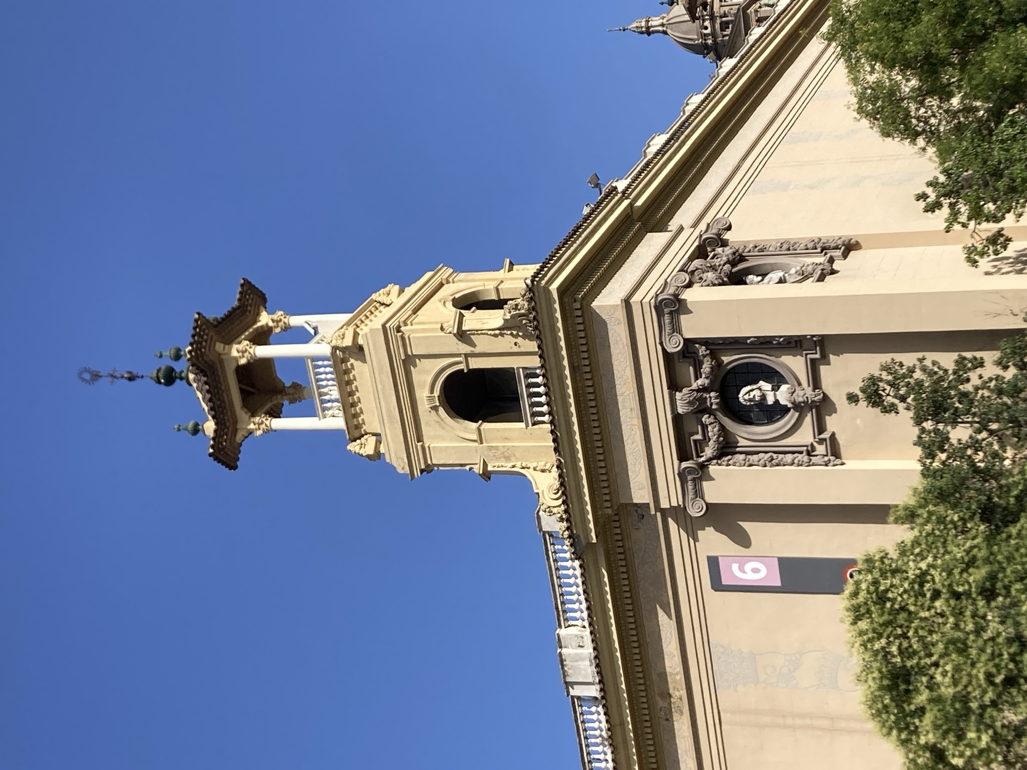 The image shows a corner of a building in Barcelona, Spain, under a clear blue sky. The building is made of a light-colored material, possibly stucco, and features classical architectural elements such as columns, moldings, and decorative reliefs. A prominent feature is a multi-tiered tower topped with a decorative spire. The tower has arched openings and a balcony. A sign with the number "6" in pink against a dark background is visible on the side of the building. Green trees partially obscure the lower part of the building. In the background, another building with a dome can be seen. The scene appears to be a street-level view of the building's exterior, possibly in a public square or along a prominent avenue.