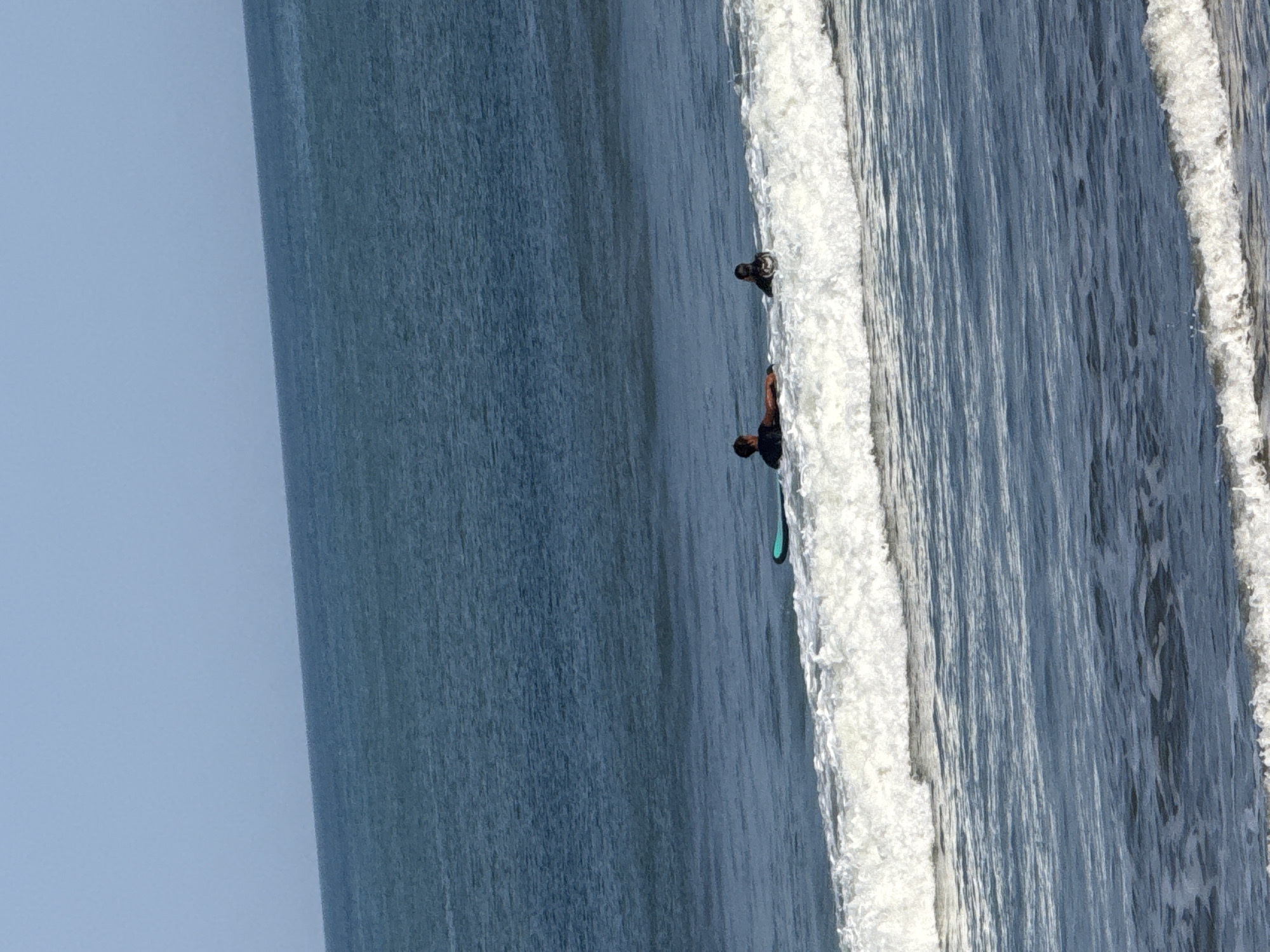 In Kuta, Indonesia, two individuals are in the ocean on surfboards. One person is lying prone on a teal surfboard, while the other sits on a black surfboard. Both are positioned in the water near a breaking wave, which is white and foamy in the foreground. The ocean is a deep blue, transitioning to a lighter blue towards the horizon, which is a clean line against a pale blue sky.