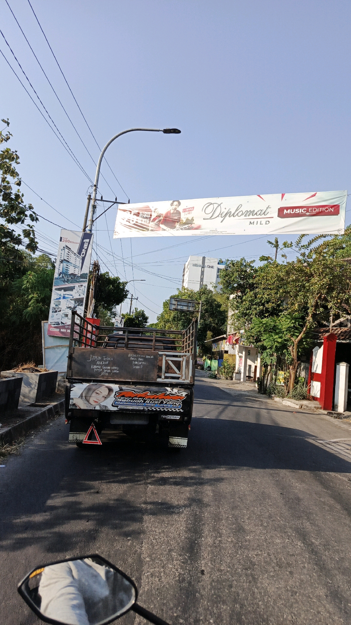 A rear-view of a pickup truck is shown driving down a street in Ngadirejo, Indonesia. The truck bed is empty, and it is adorned with various stickers and text. One sticker features a young woman with blonde hair, while another includes a phrase in Indonesian that translates to "Don't be afraid of the tough times, because success is what comes from hard work." The truck is also equipped with a red triangular reflective marker on its rear. Overhead, a large banner hangs across the street advertising "Diplomat MILD MUSIC EDITION." Several utility poles and power lines are visible, along with trees and buildings lining the street. The sky is clear and blue, indicating a sunny day. The image appears to be taken from another vehicle, as a motorcycle mirror is visible in the bottom left corner.