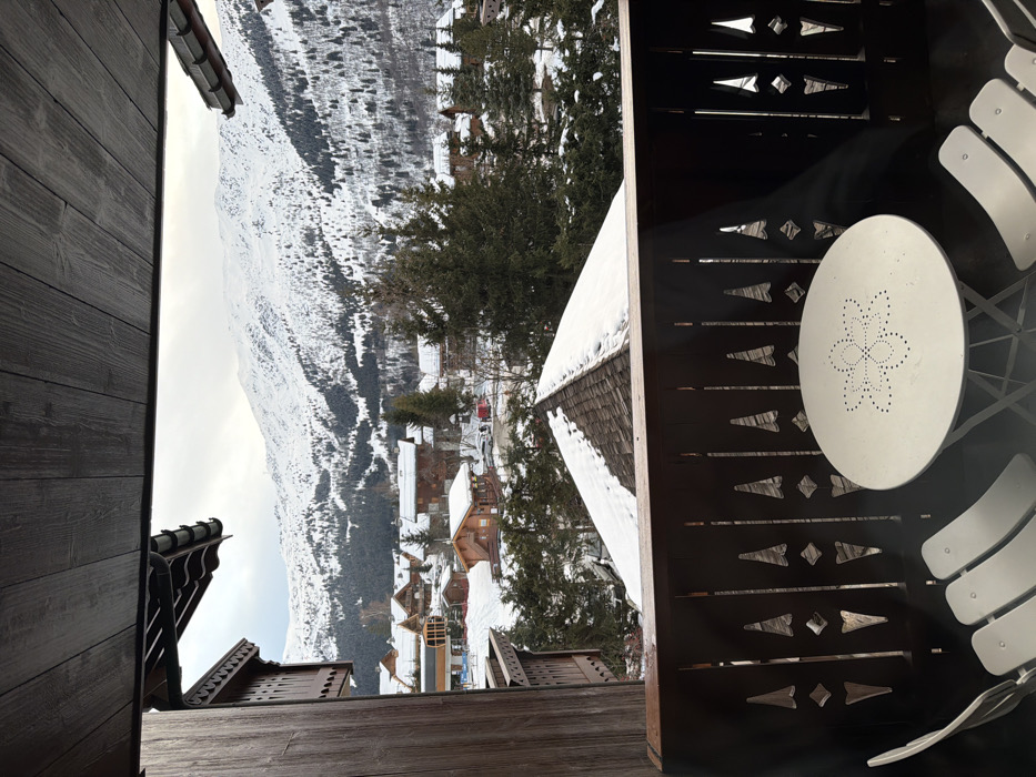 A snowy mountain vista is seen from a balcony. A white table with a flower design sits on the balcony, surrounded by white chairs. A dark brown railing with decorative cutouts encloses the space. Snow-covered rooftops of chalets and tall pine trees dot the landscape below the mountains. The sky is overcast. Wood paneling of the building is visible on the left side of the frame.