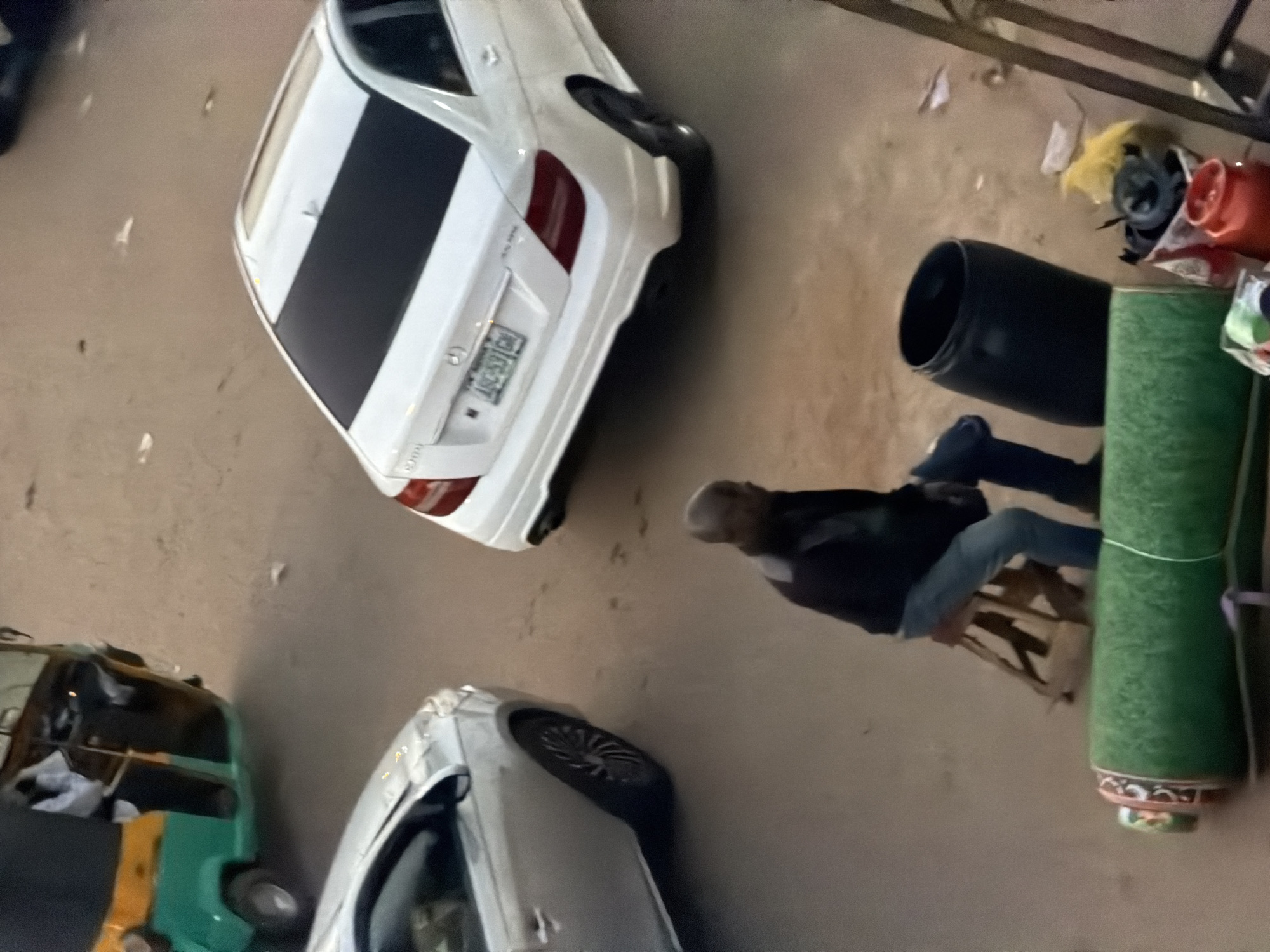 In Bukuru, Nigeria, a man sits on a wooden stool beside a large black barrel. In front of the man, a rolled-up green mat lies on the ground. To the man's right, a white Mercedes-Benz sedan is parked. To the left of the man, a silver car is parked. In the background, a three-wheeled auto-rickshaw is visible. The scene appears to be a street with a dusty surface.
