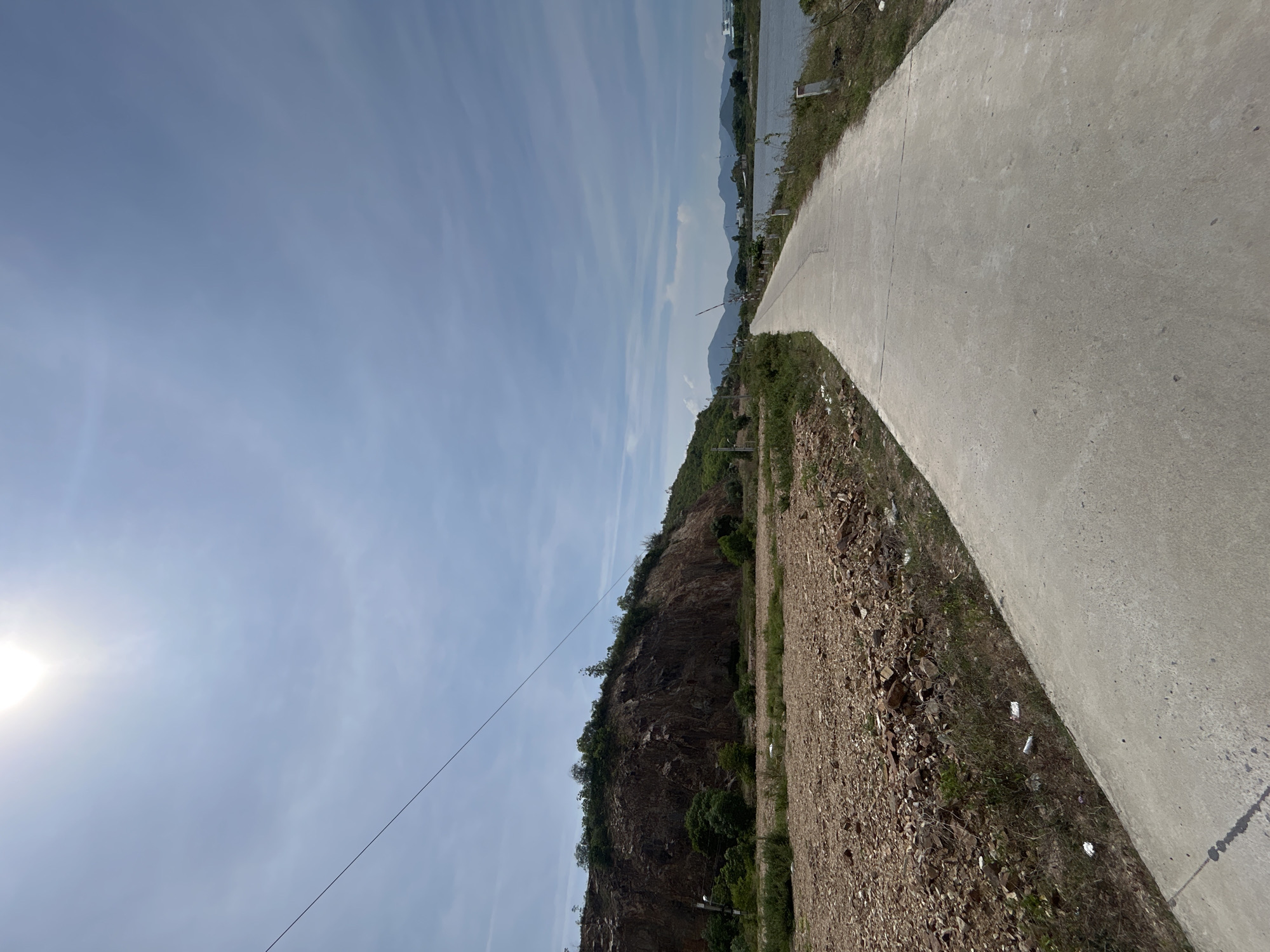 The image depicts a paved road curving towards a body of water under a bright, partly cloudy sky. To the left of the road is a steep, rocky hillside covered with sparse vegetation and trees. The ground in the foreground to the left of the road is covered with loose stones and dirt. The road itself is concrete and shows faint expansion joints. In the distance, mountains are visible across the water. The overall scene suggests a rural or developing area, possibly on the outskirts of a city like Nam Nha Trang Ward, Vietnam. There are no people or animals visible in the image. The environment is natural with man-made elements such as the road and a distant structure that could be a building. A power line stretches across the sky from the upper left to the middle right.