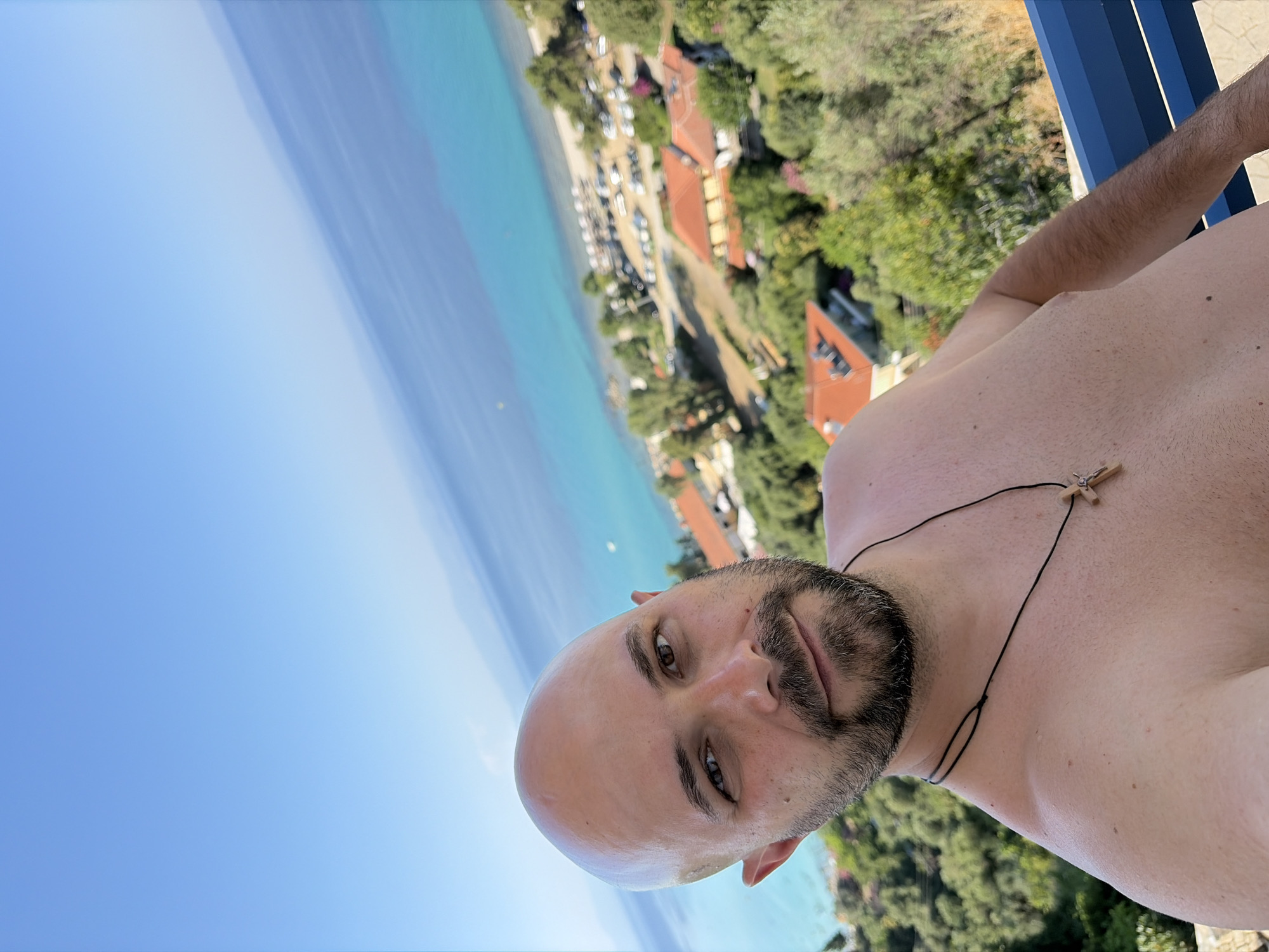 The image is a selfie taken by a bald, bearded man wearing a necklace with a wooden cross. He is shirtless and appears to be on a balcony or terrace overlooking a coastal town in the Kassandra Municipal Unit, Greece. The background shows a turquoise sea, a sandy beach with beach chairs and umbrellas, parked cars, and houses with red-tiled roofs nestled among green trees. The sky is a clear, bright blue. The man is looking directly at the camera with a slight smile. The overall impression is one of a sunny vacation in a Mediterranean location.