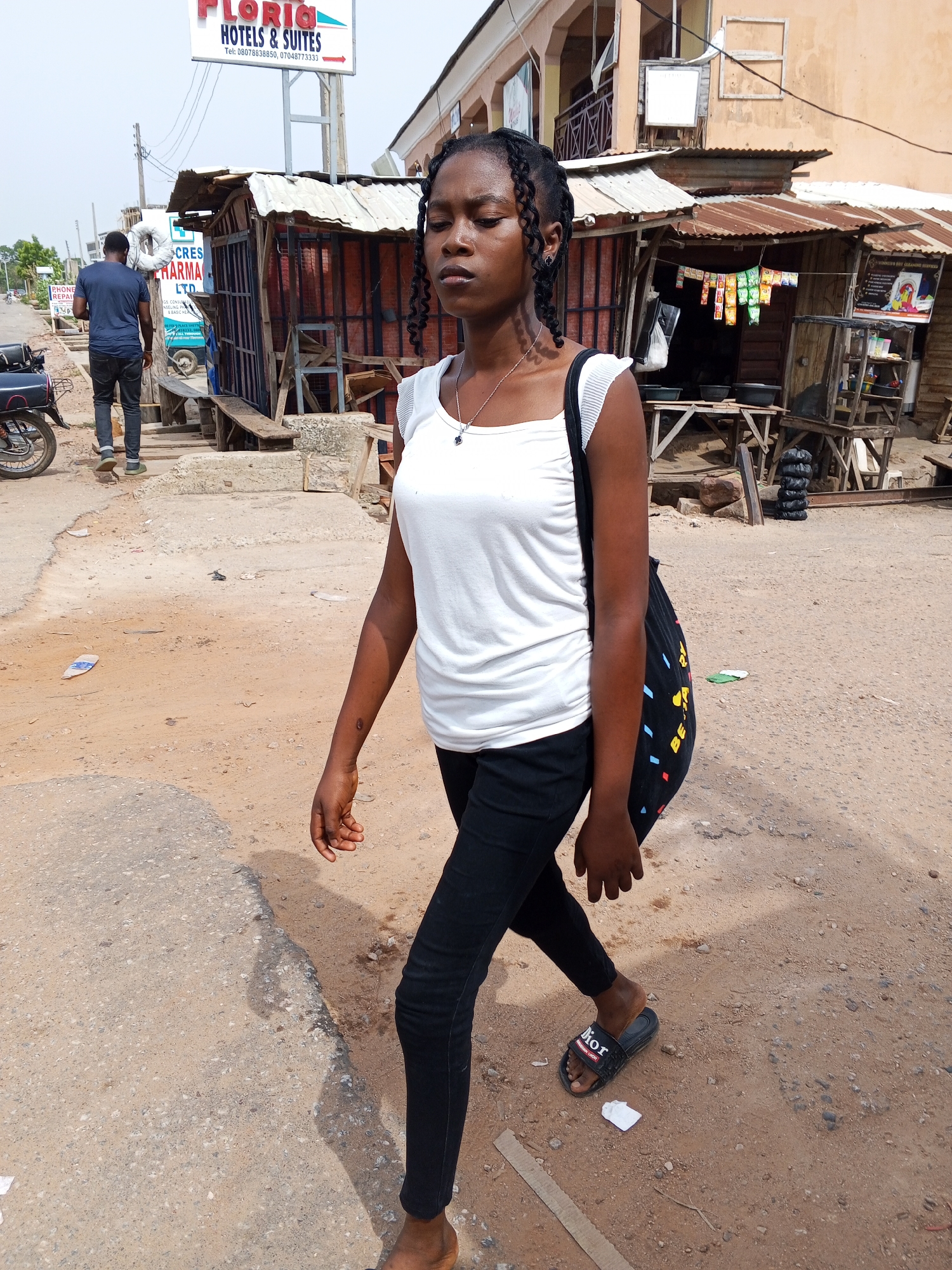 A young, dark-skinned woman is walking toward the camera in Adankolo, Nigeria. She wears a white top, black pants, and Dior-branded slide sandals. A black shoulder bag hangs from her left shoulder. Her hair is styled in braids.
In the background, a man in a blue shirt and dark pants is walking away. A motorcycle is parked nearby. There are makeshift commercial structures, including one advertising "Cres Pharma LTD." In the upper part of the frame, a sign reads "Floria Hotels & Suites." The ground is uneven, with a mix of dirt and asphalt.