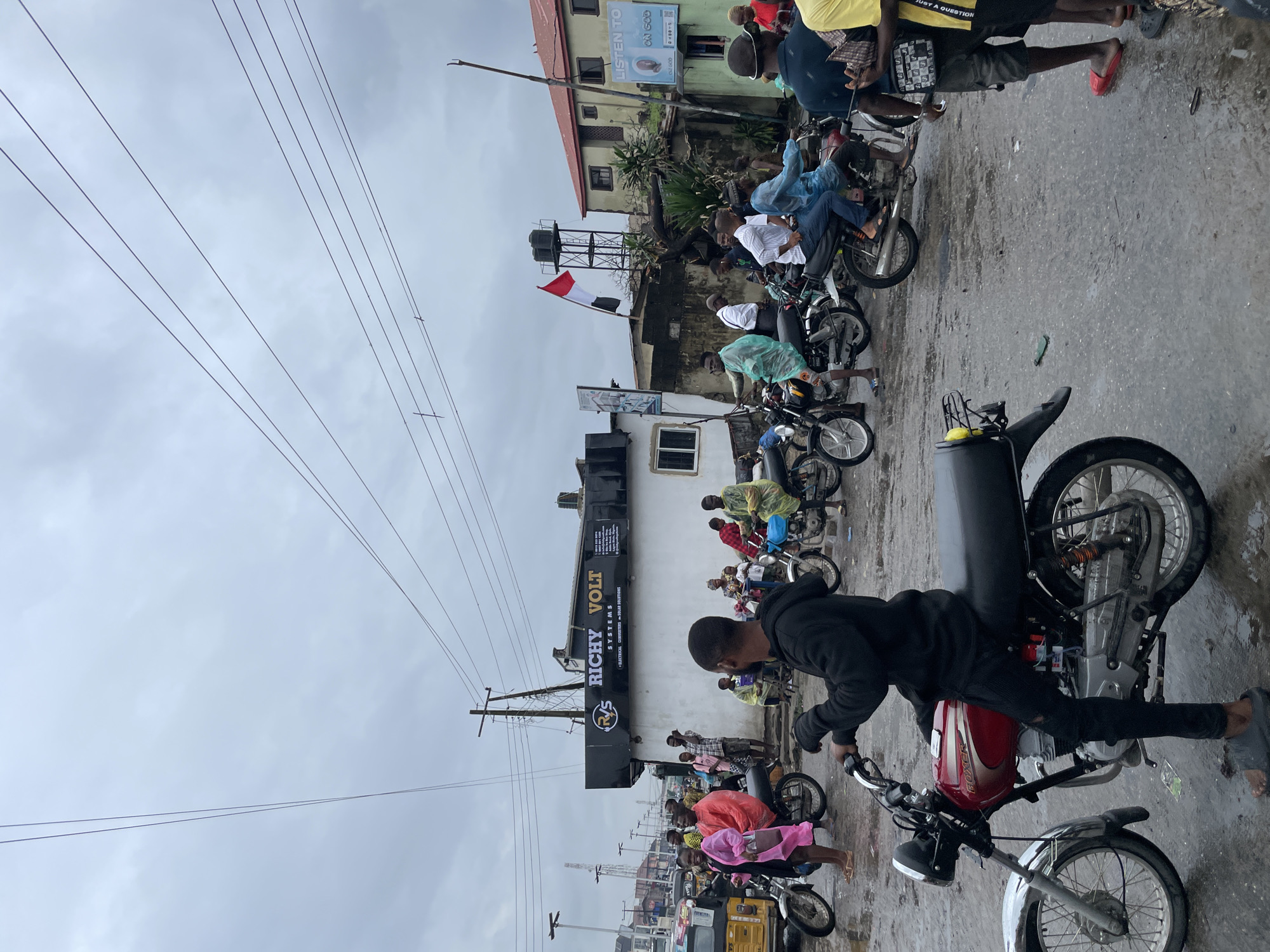 The image depicts a busy street scene in Ado, Nigeria, under an overcast sky, with a group of people gathered around motorcycles. The ground is wet, suggesting recent rain. In the foreground, a person on a red motorcycle wearing a black hoodie is visible from the back. Behind them, several other motorcycles are parked or in motion, carrying passengers. Some individuals are wearing colorful raincoats or ponchos, indicating the weather. A building with signage for "RICHY VOLT SYSTEMS" is prominently displayed. Further in the background, other buildings and street infrastructure like utility poles and power lines are visible. A flag with red and white horizontal stripes is flying on a pole. The overall impression is one of a bustling urban environment with a focus on motorcycle transportation.