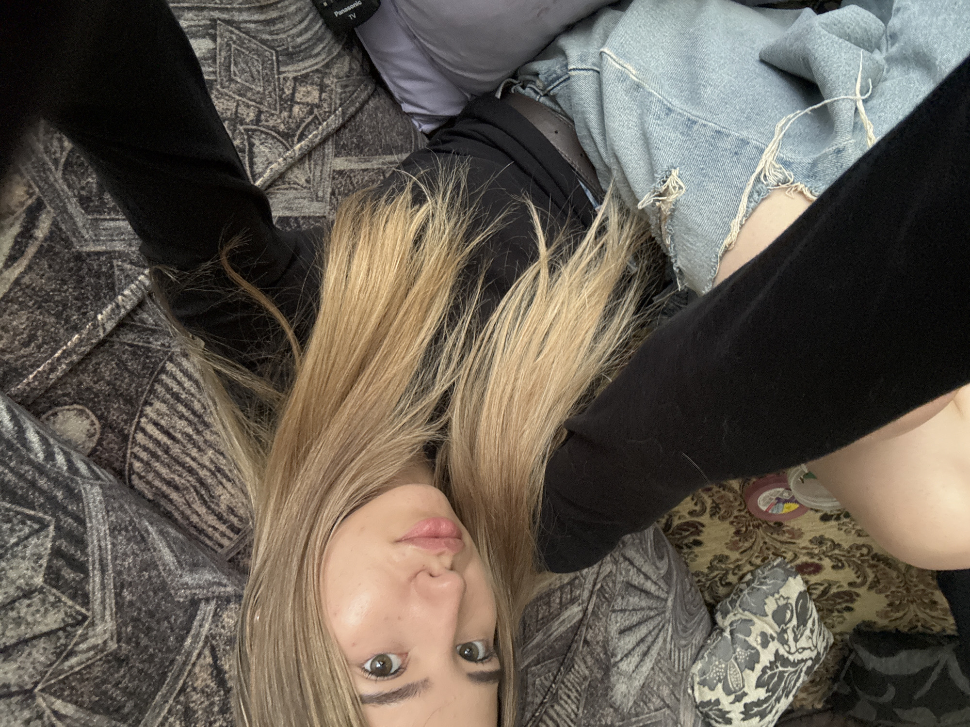 A young woman with blonde hair and green eyes is lying down and taking a selfie. She is wearing a black long-sleeved shirt and ripped denim shorts. The background appears to be a sofa with patterned cushions and blankets. The lighting suggests it is indoors, possibly in a home environment. There are no specific details in the image that can be used to determine her location in Noviy, Russia.